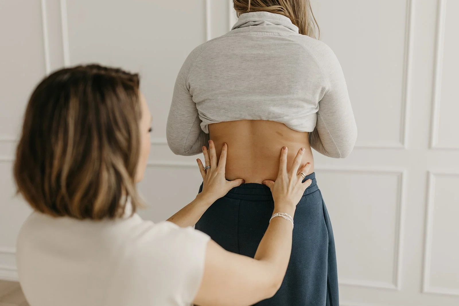 Woman with a grey cropped hoodie with her back turned, having her waist measured by another woman with shoulder-length brown hair, white top, in a room with white walls.