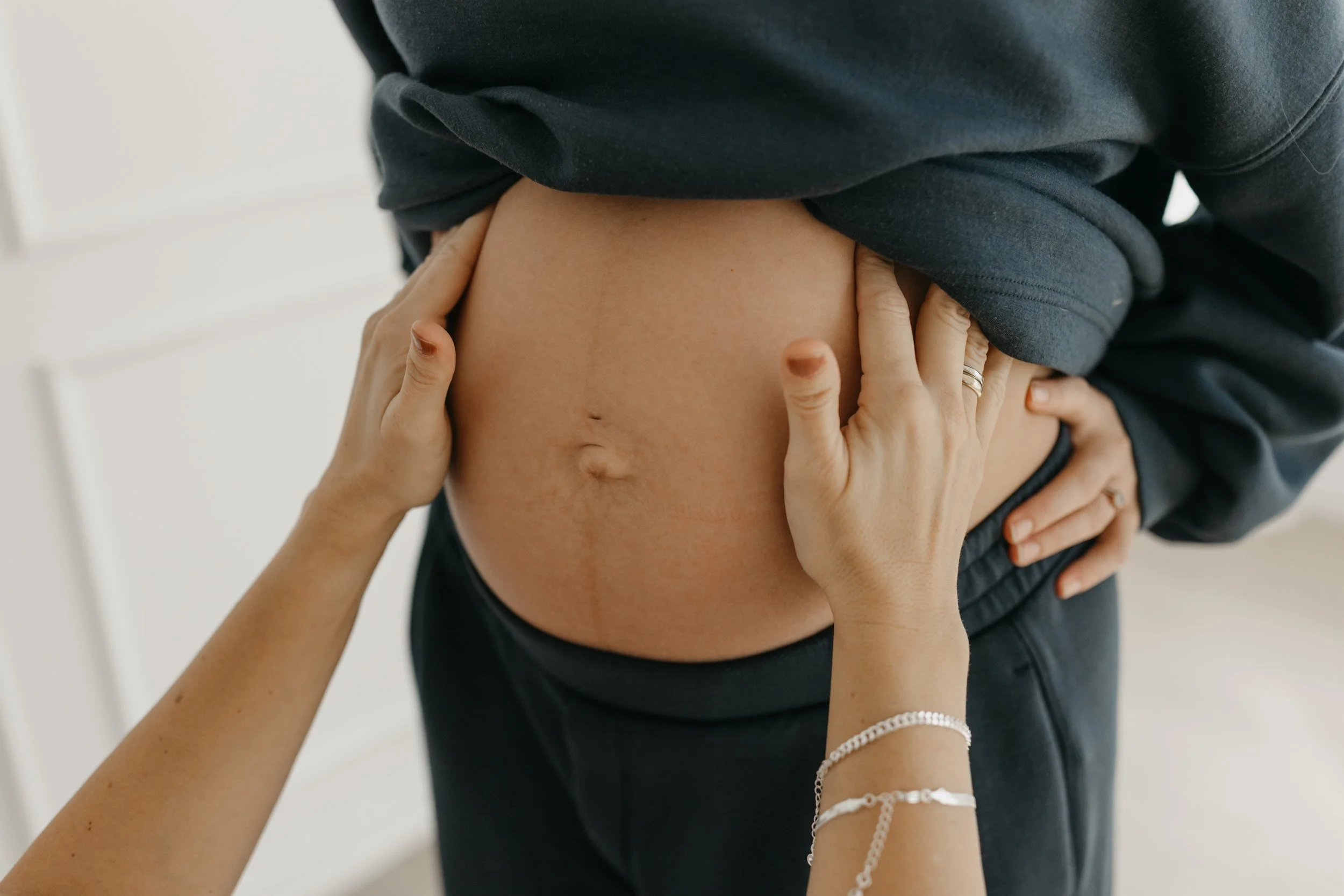Person lifting shirt to reveal their bare pregnant belly while another person touches their stomach.