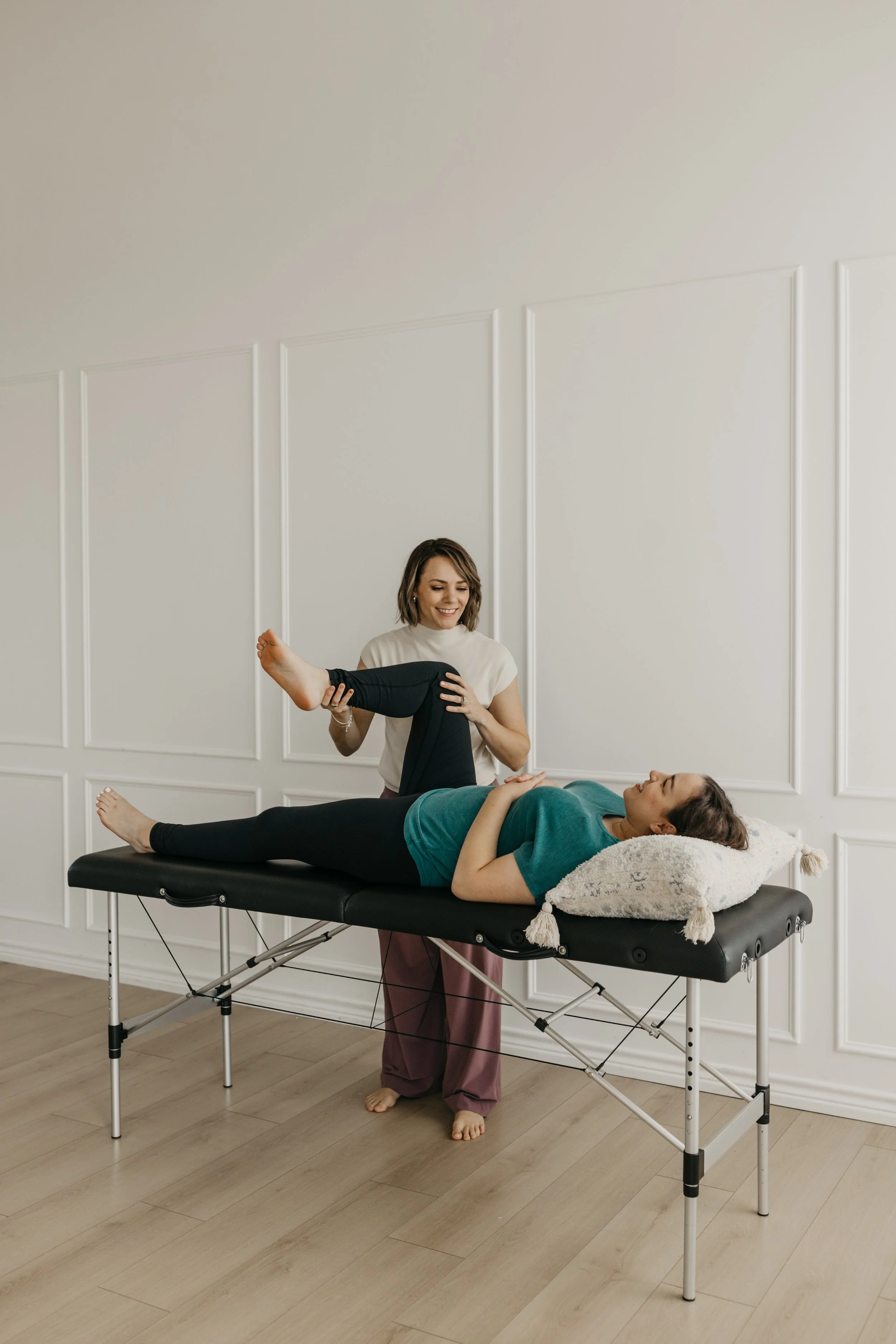 A woman lying on a massage table receiving physical therapy while a therapist assists her with leg exercises in a bright, minimalistic room.