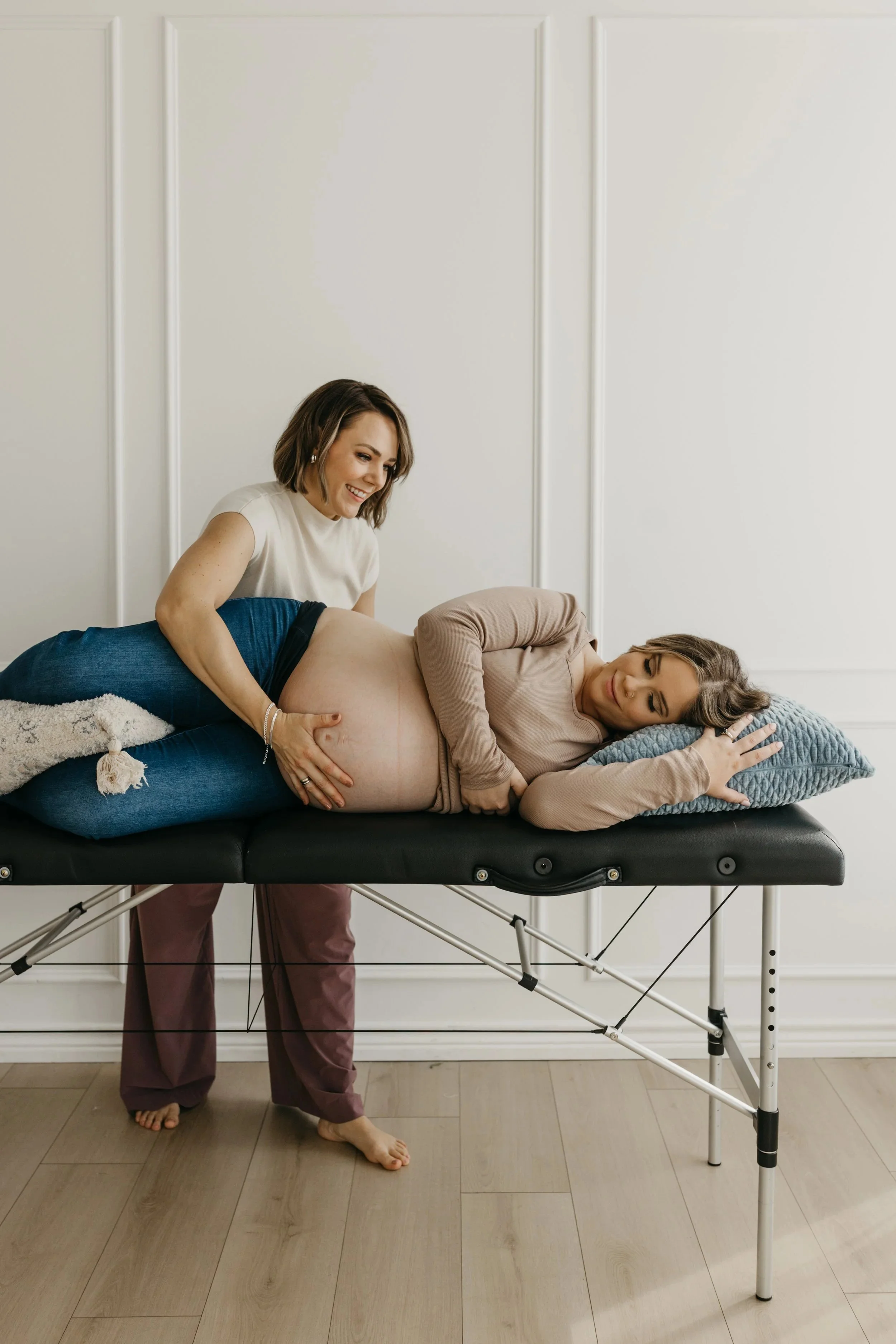 Pregnant woman lying on a massage table, receiving a prenatal massage from another woman in a soft beige top, in a bright, neutral room.