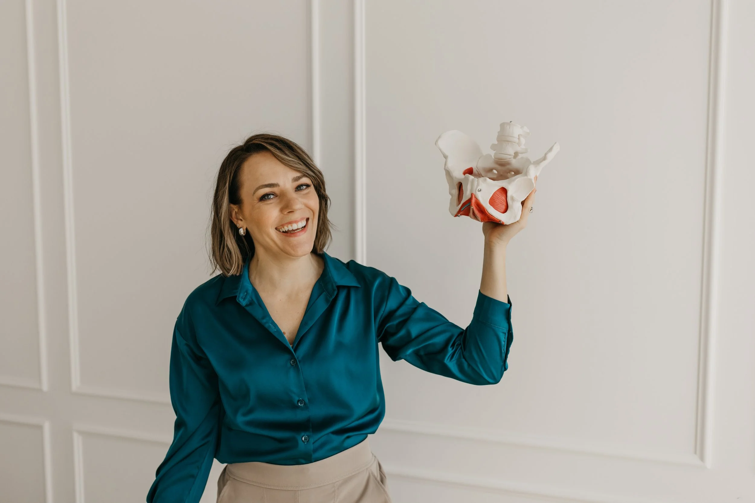 A woman with short brown hair, smiling, dressed in a teal blouse and beige pants, holding a pelvic bone model, standing against a plain wall.