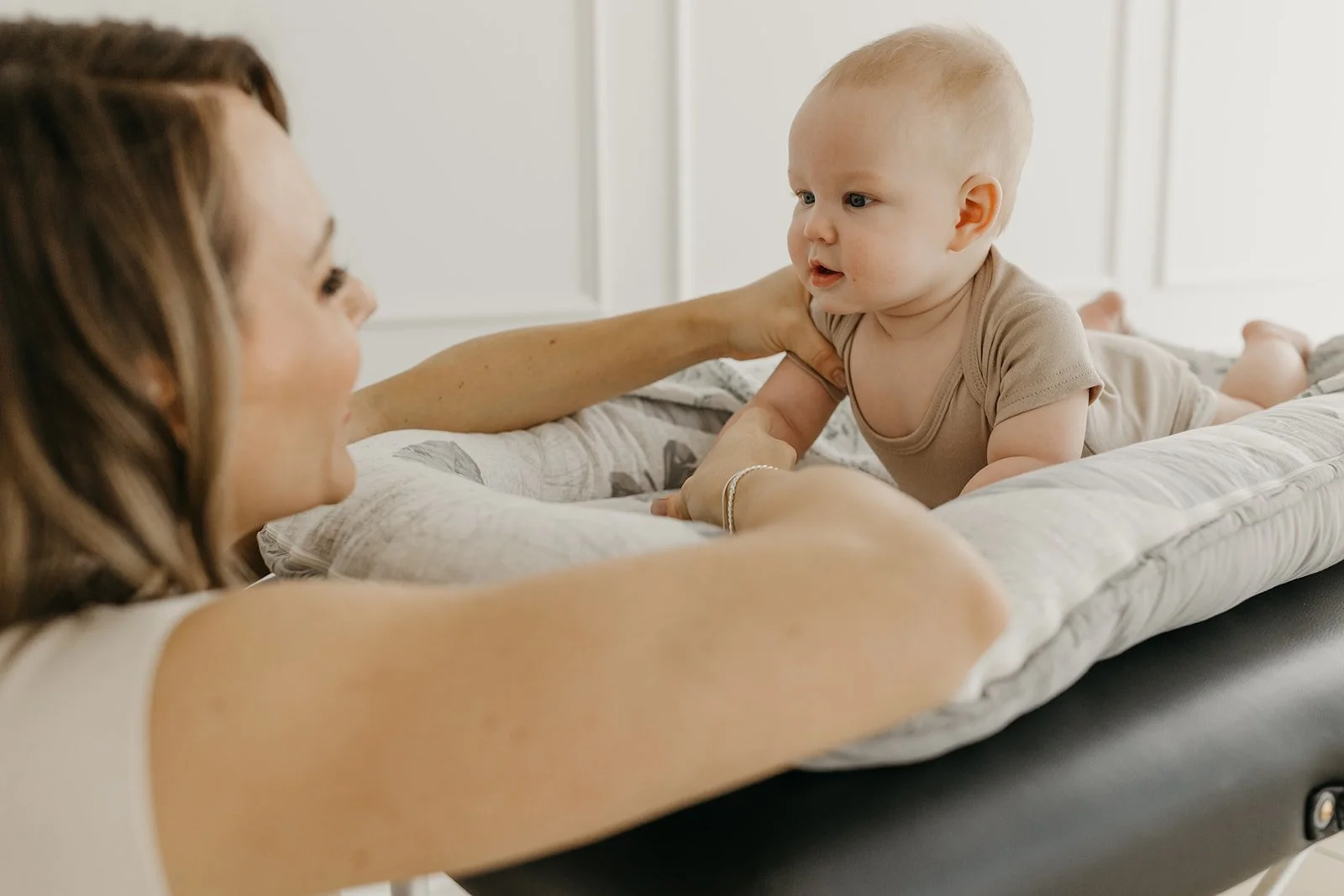 A woman and a baby engaging in play on an elevated changing table, with the woman lying on her side and the baby on stomach, facing each other, in a bright room.