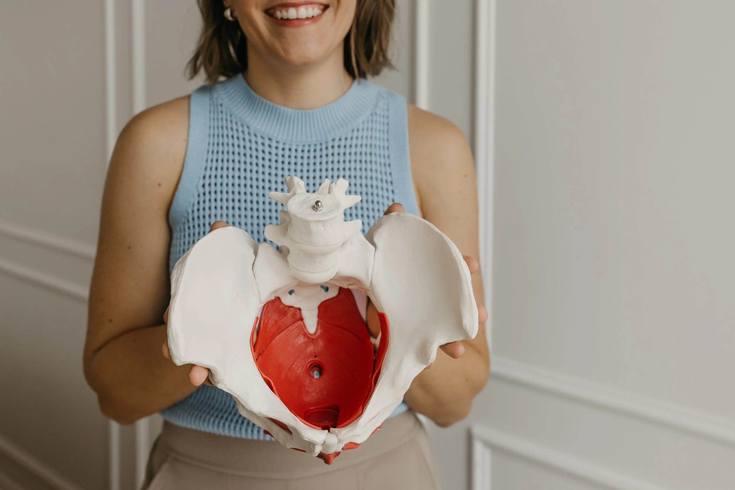 A woman holding a detailed anatomical model of the human pelvis and lower spine, smiling in a professional setting with white paneled walls.