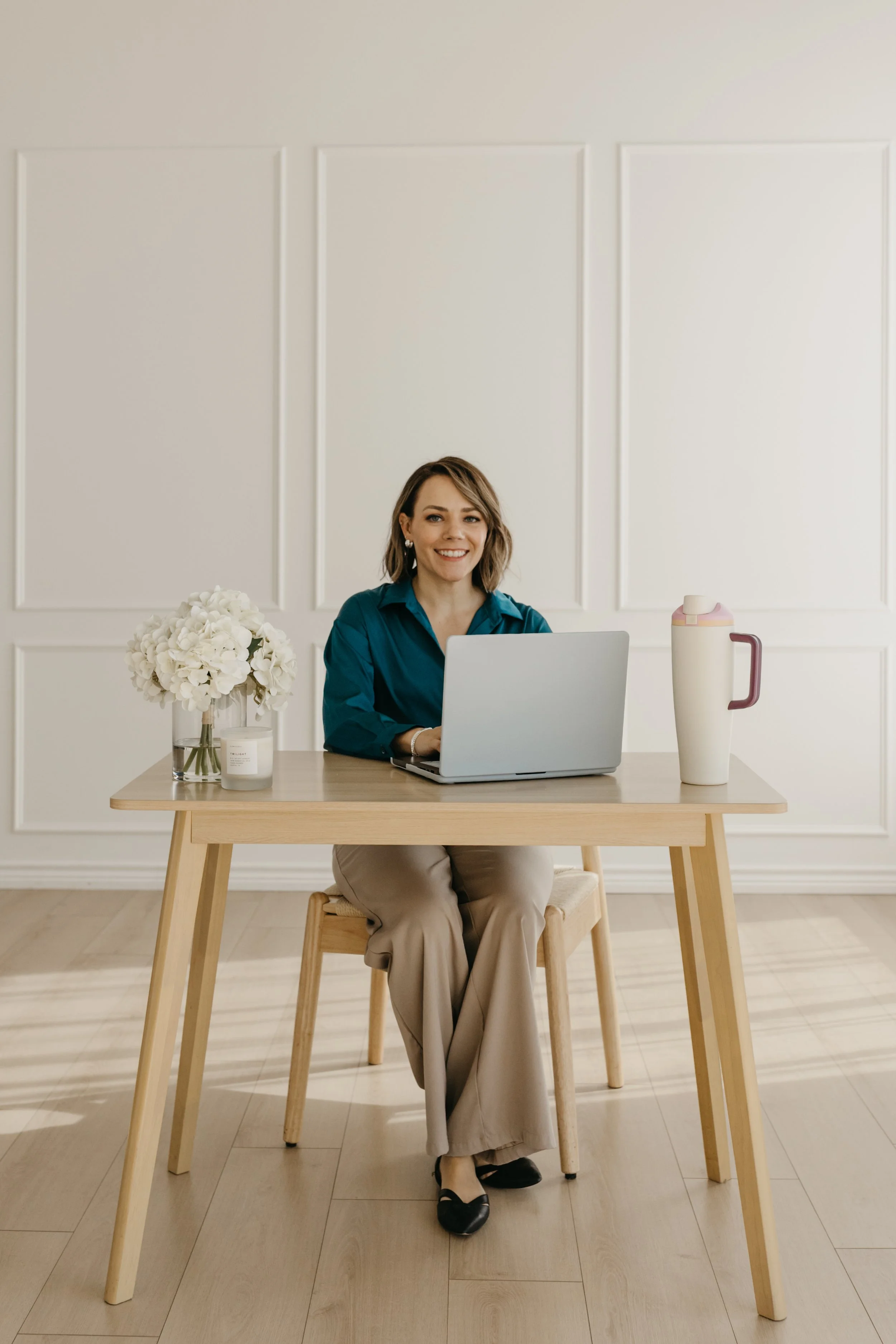 A woman sitting at a wooden table with a laptop, flowers, a candle, and a water jug in a bright, minimalist room.