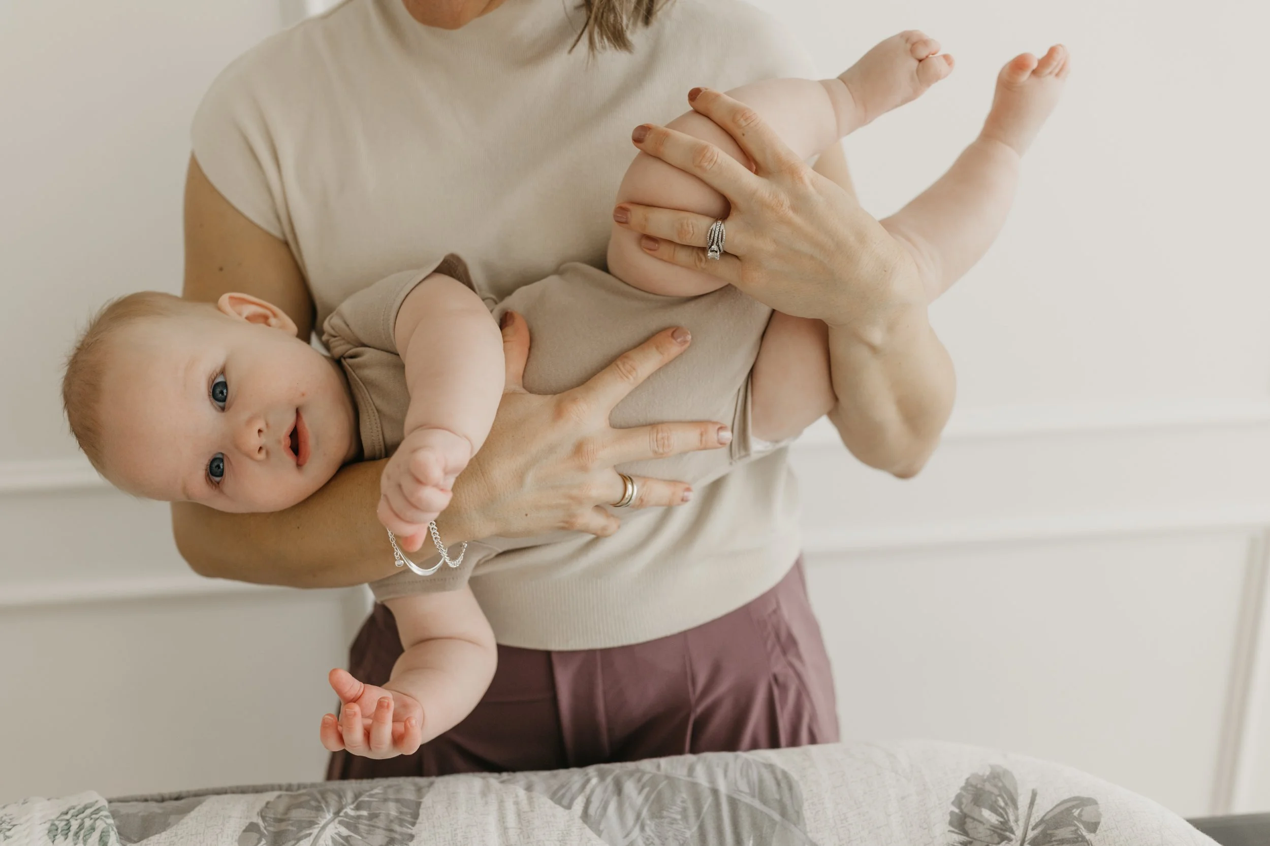 A woman holding a baby horizontally, supporting the baby's back and legs, with the baby looking at the camera. The woman is wearing rings and a beige shirt, and the baby is dressed in a beige onesie. The background is a neutral-colored wall.