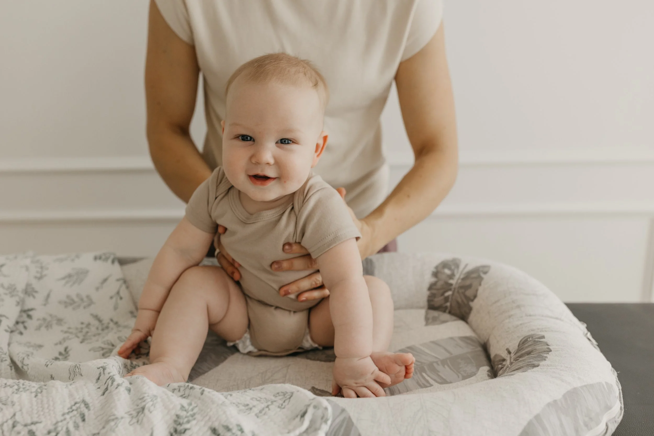 A smiling baby with light hair, sitting on a changing pad, with an adult supporting behind.