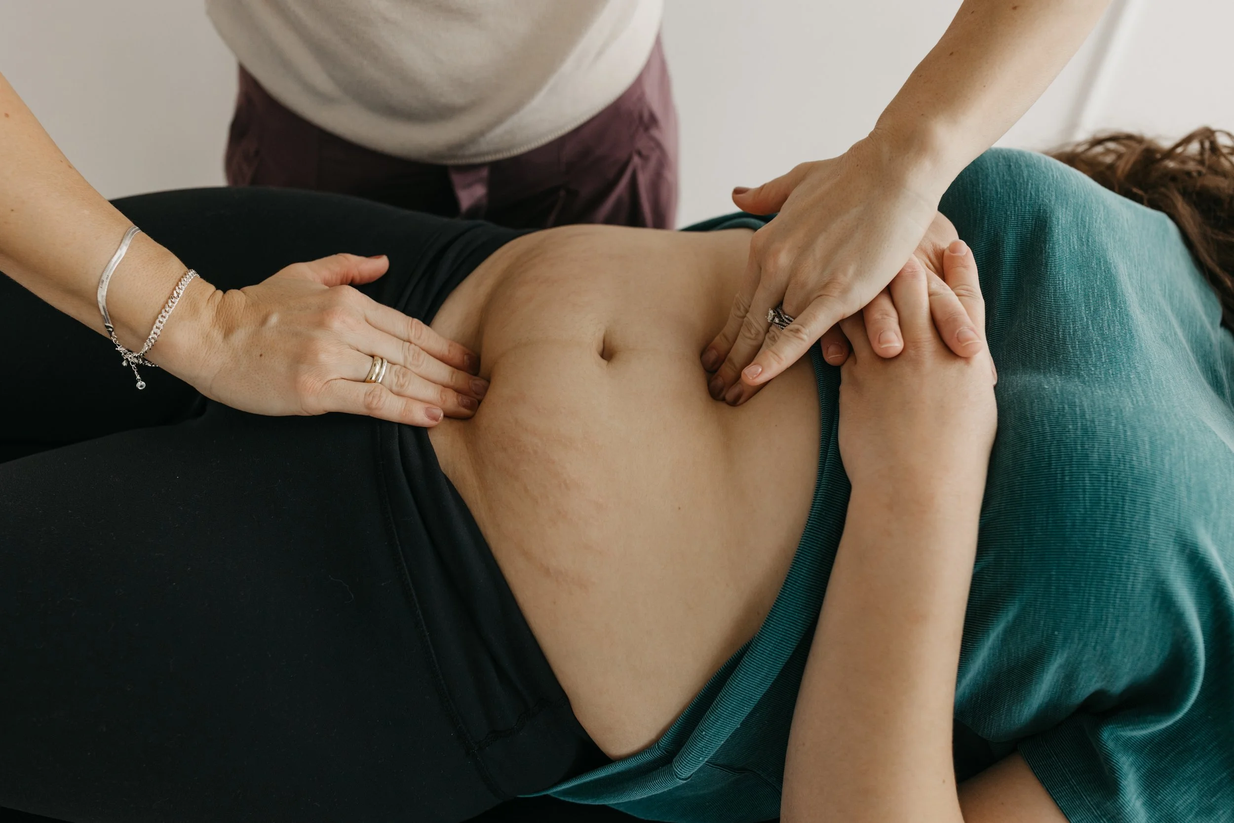 A person receiving chiropractic or massage therapy, lying on a treatment table while a practitioner presses on their abdomen, with another person's arm visible in the foreground.