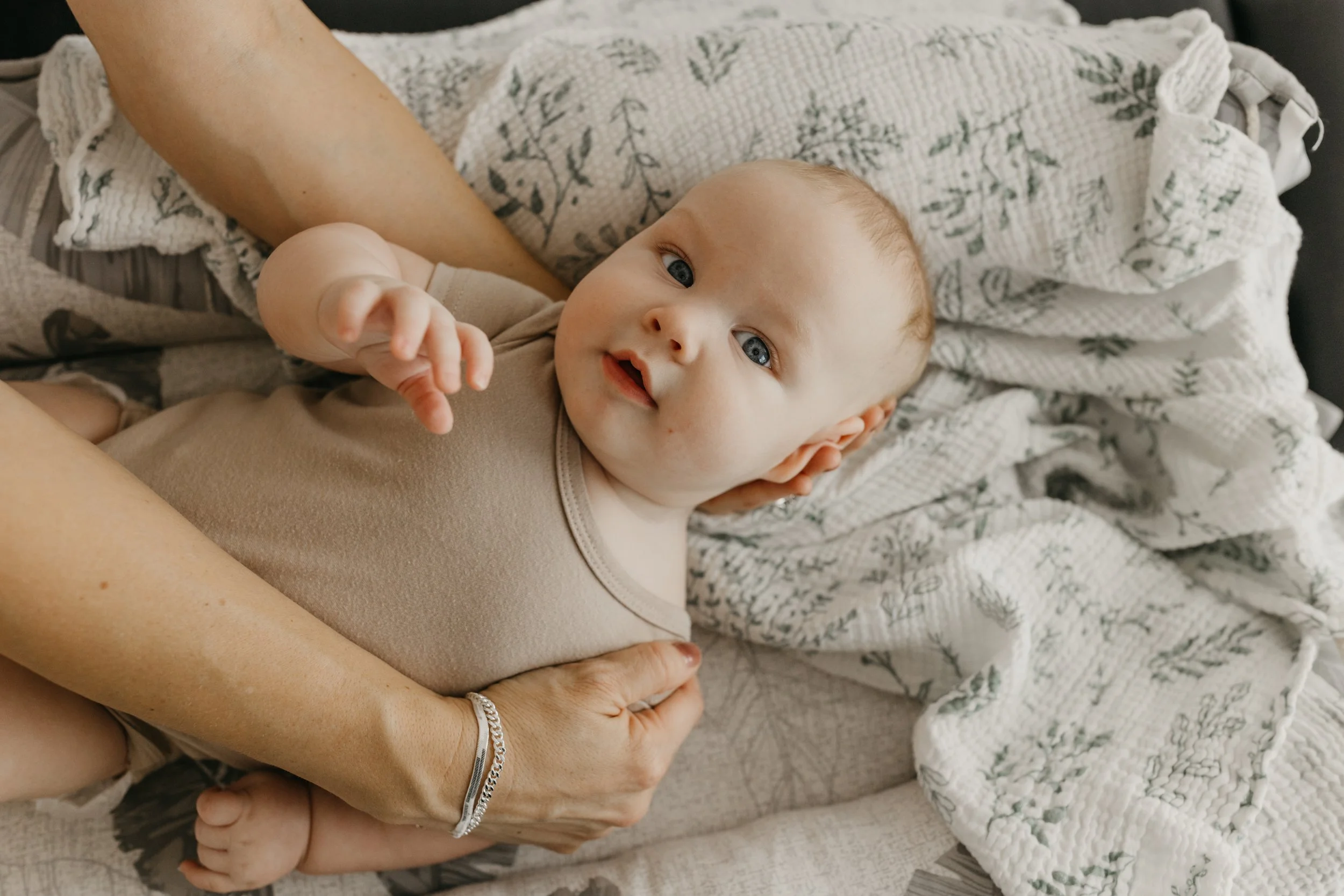 A baby with blue eyes lying on a bed with an adult's hands supporting the head and body, surrounded by white and gray patterned blankets.
