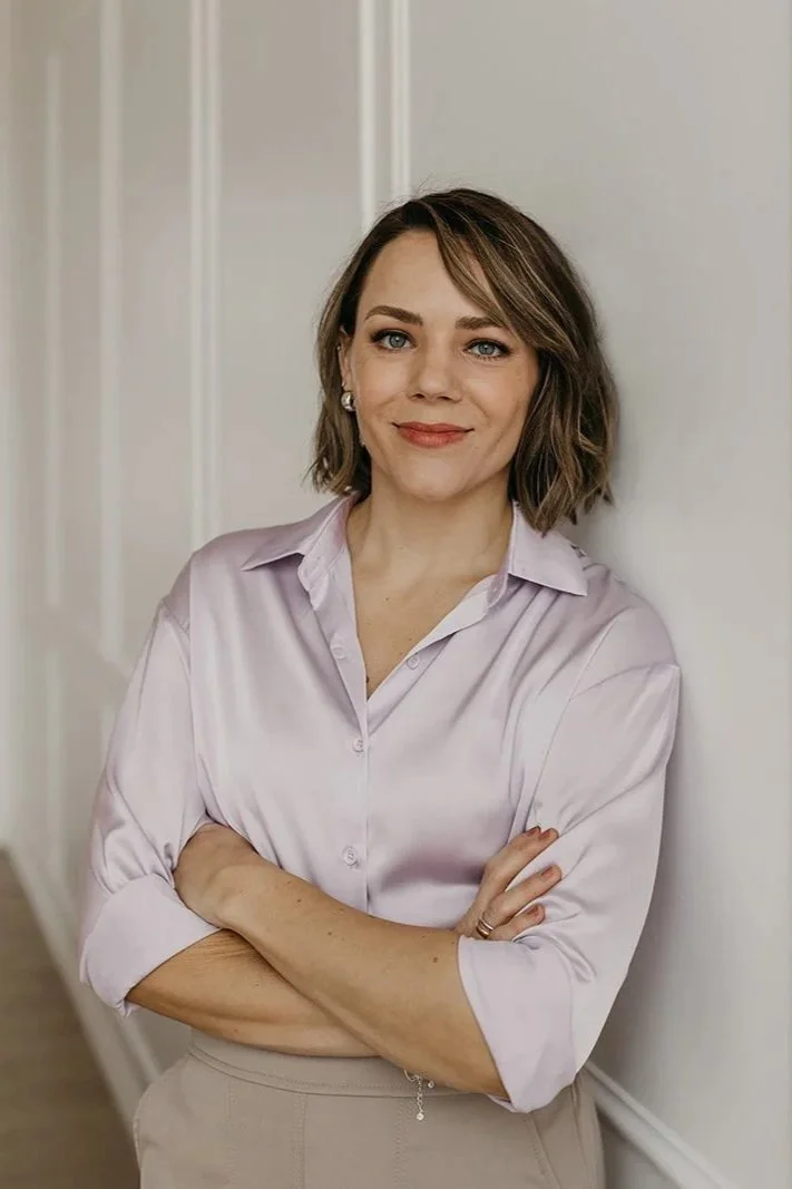 A woman with short, wavy brown hair and blue eyes, wearing a light pink satin blouse and beige pants, standing with arms crossed and smiling against a neutral background.