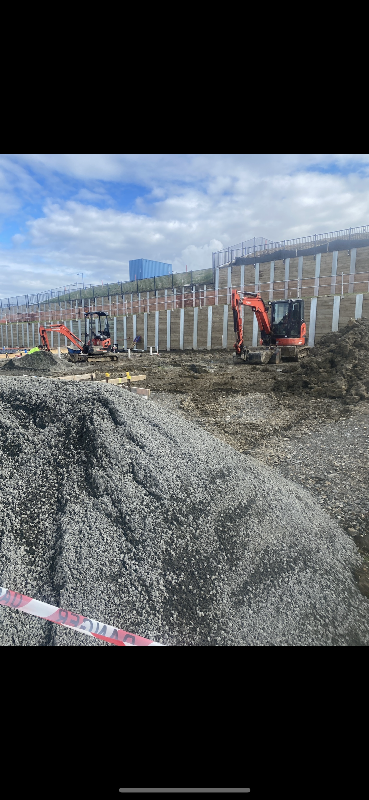 Construction site with two small excavators working on earthwork, with a gravel pile in the foreground and a retaining wall with fencing in the background.