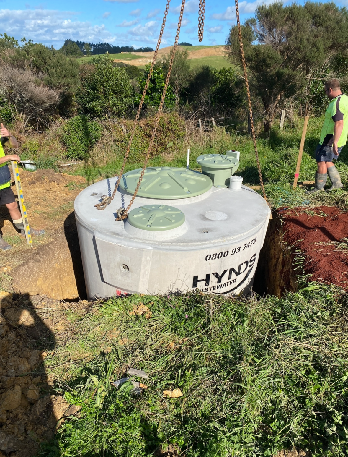 A large septic tank being installed in a rural outdoor setting with two workers nearby, surrounded by grass, bushes, and rolling hills.