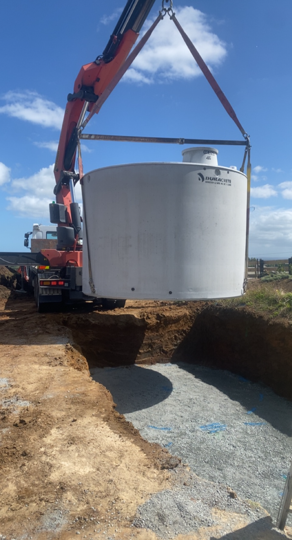 Construction crane lowering a large concrete basin into a prepared excavation site under a blue sky with scattered clouds.