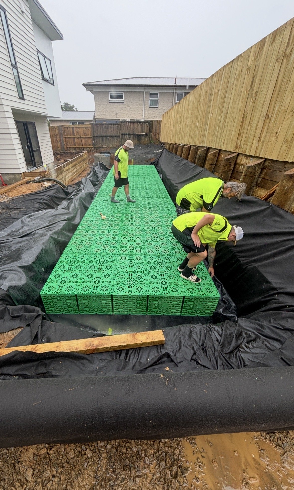 Workers in yellow safety vests install green concrete pavers on a patio. The area is prepared with a black geotextile fabric, and there is a wooden retaining wall beside the construction site.