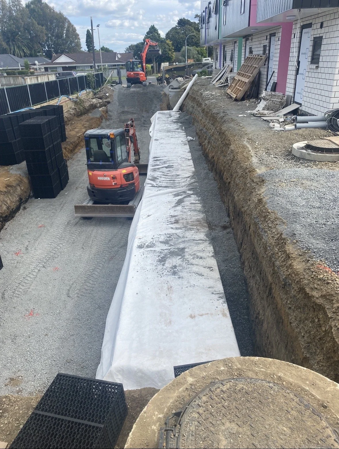 Construction site with a small excavator laying fabric in a trench, with building and materials in the background.