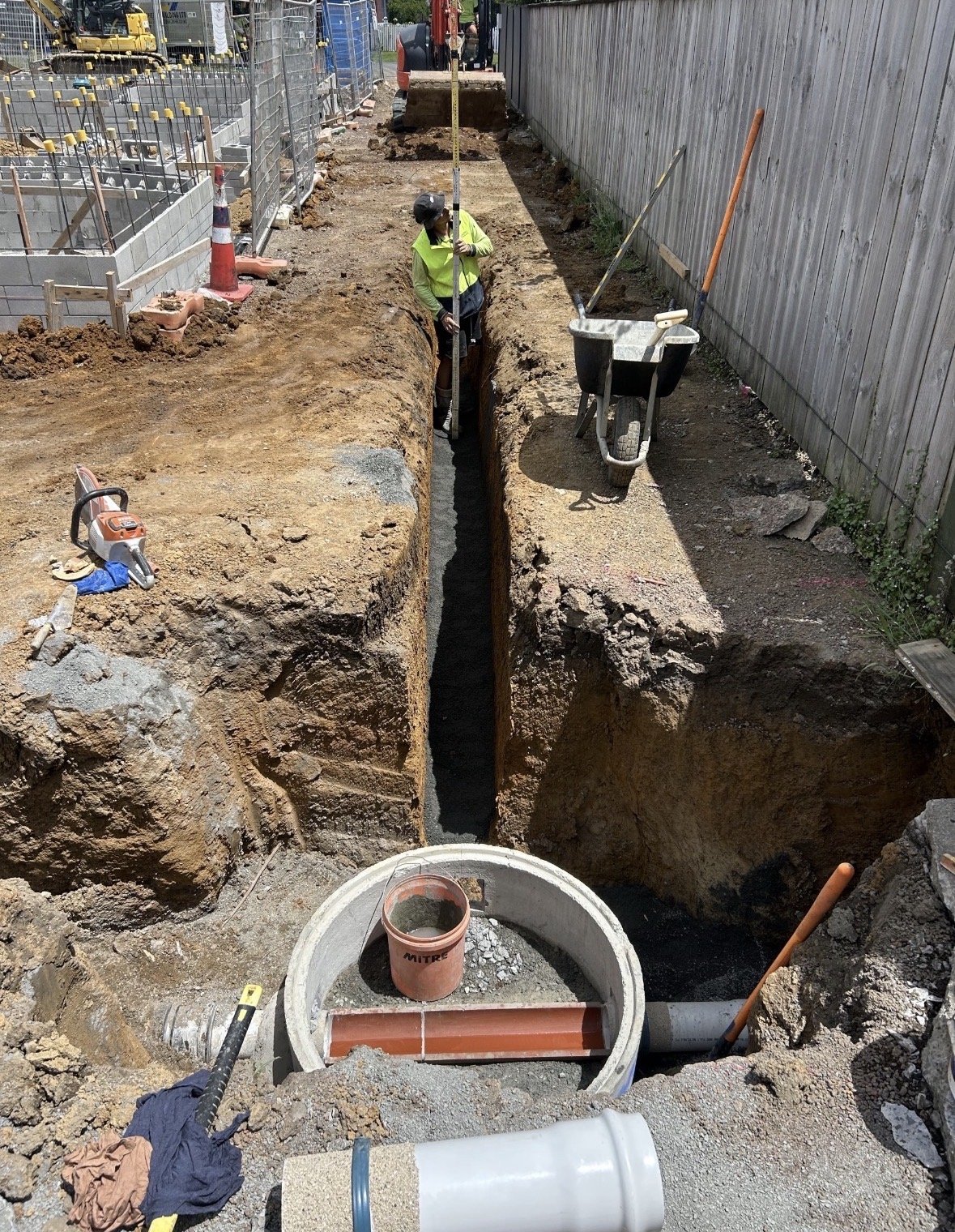 Construction workers install underground piping in a narrow trench with a wheelbarrow and construction tools nearby.