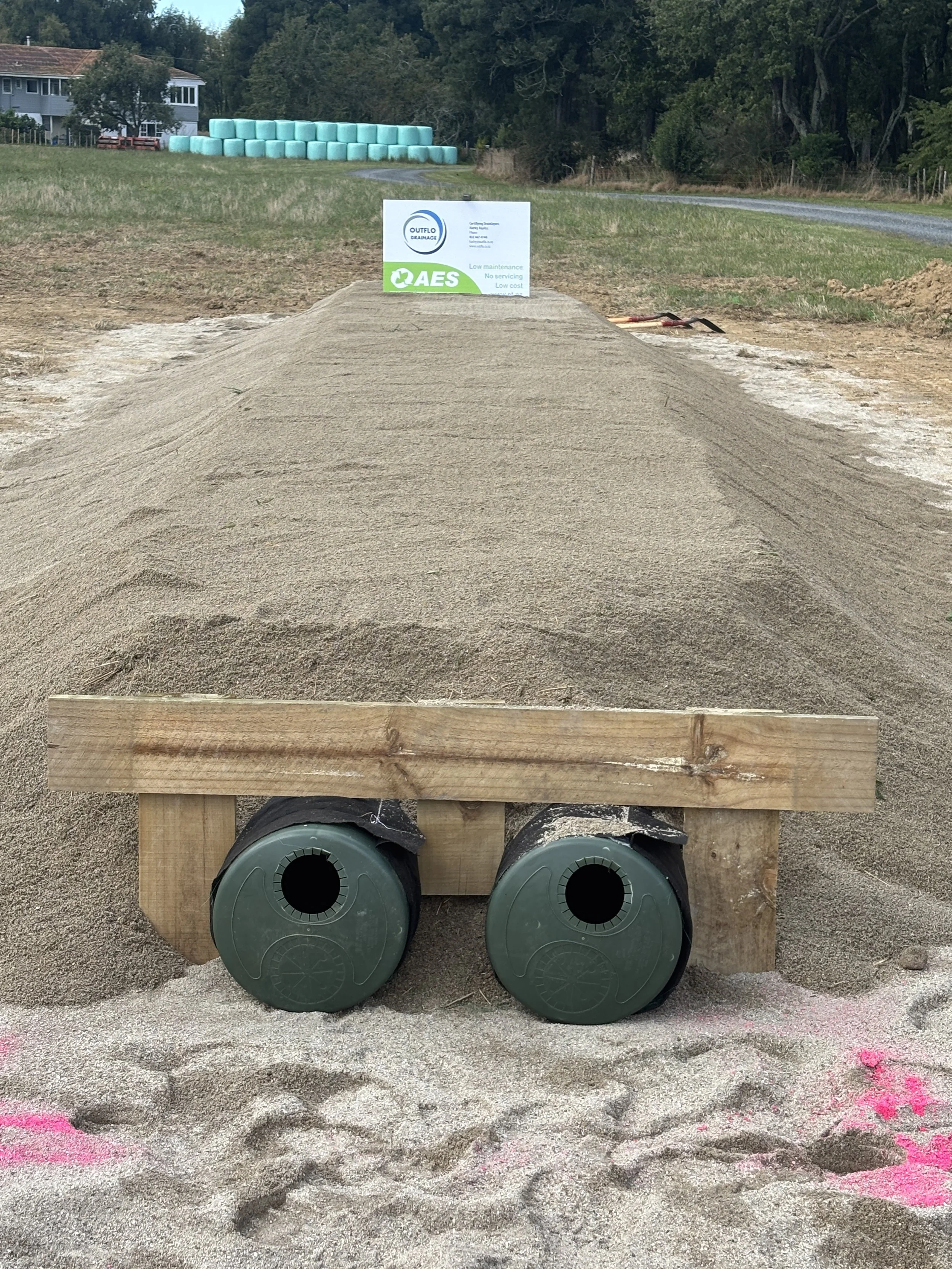 Sand mound with two black drainage pipes at the base, a wooden board on top, and a sign for outlet drainage in the background, with hay bales and trees behind.