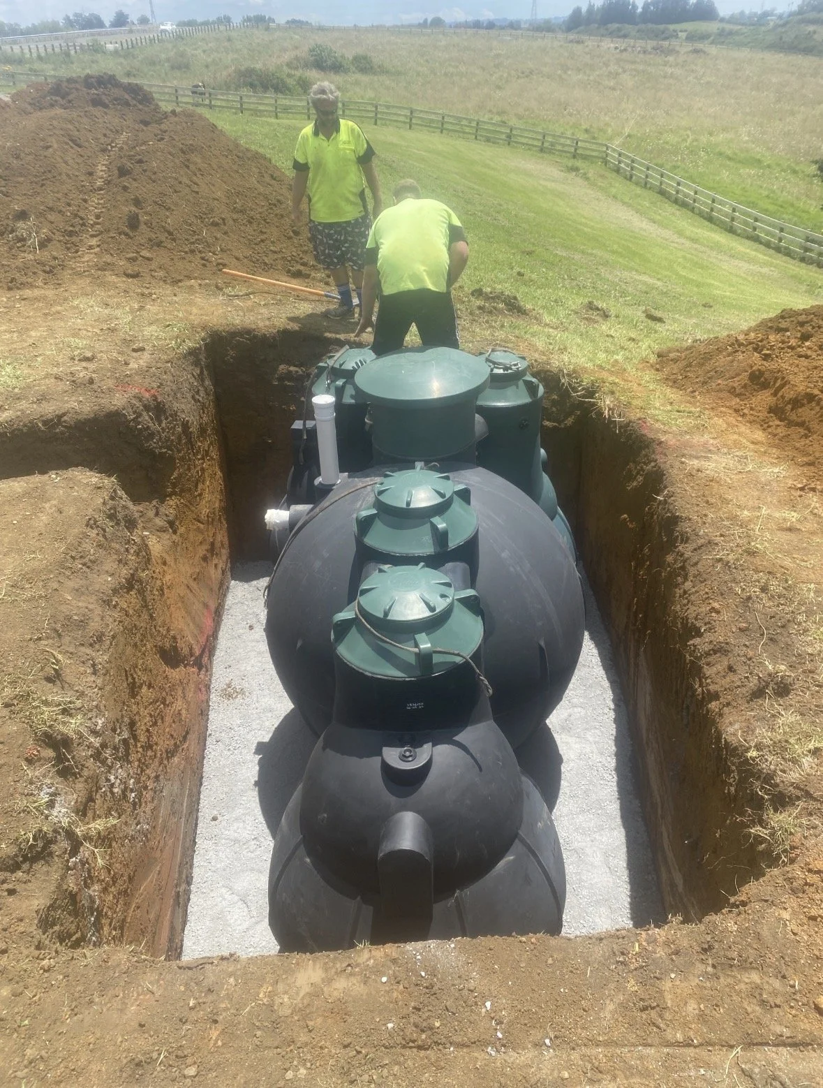 Three people working in a trench with a large underground water tank in a grassy outdoor area.