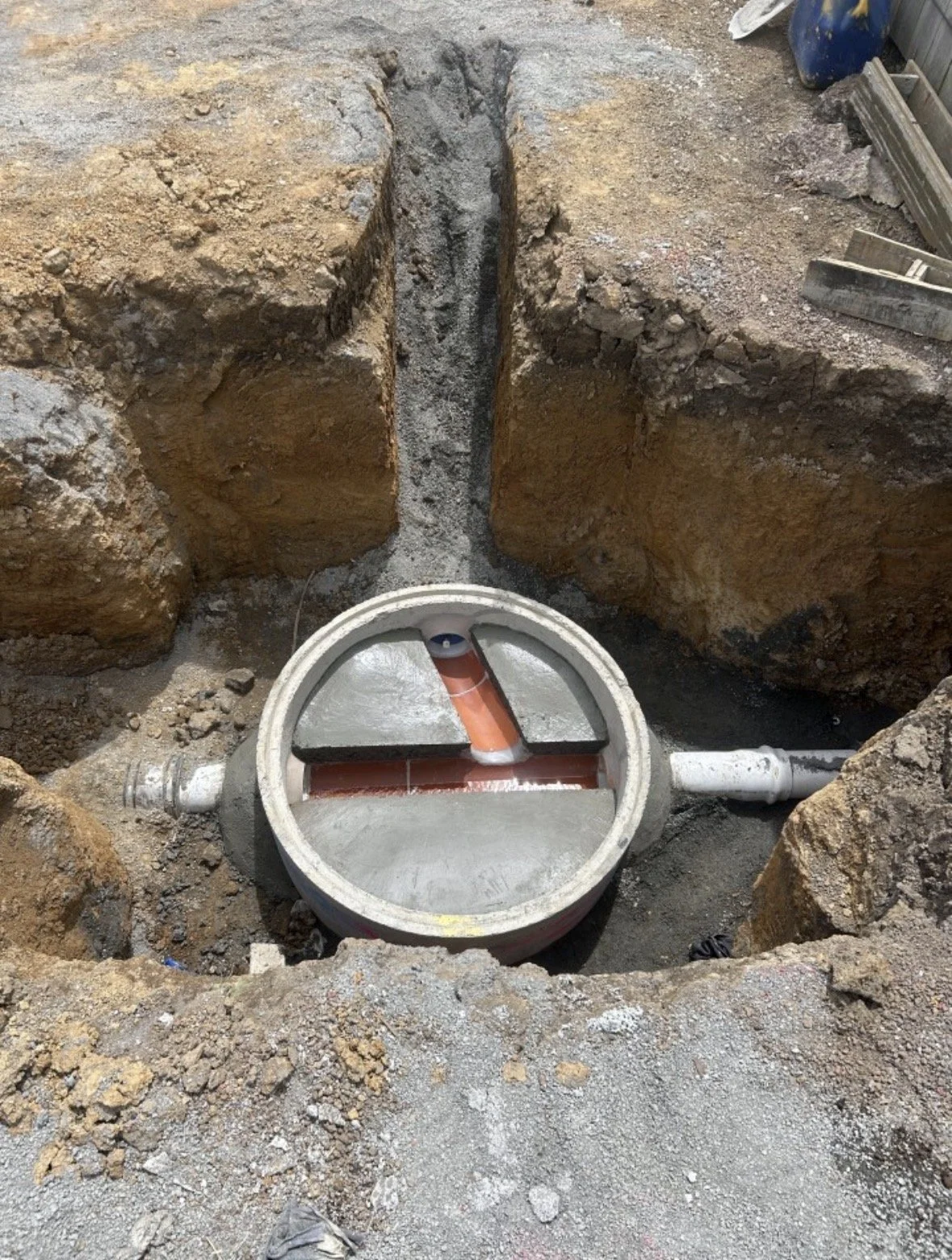 Construction site with an underground drainage or sewer system with a large pipe fitting and red pipe inside the opening, surrounded by dirt and soil.