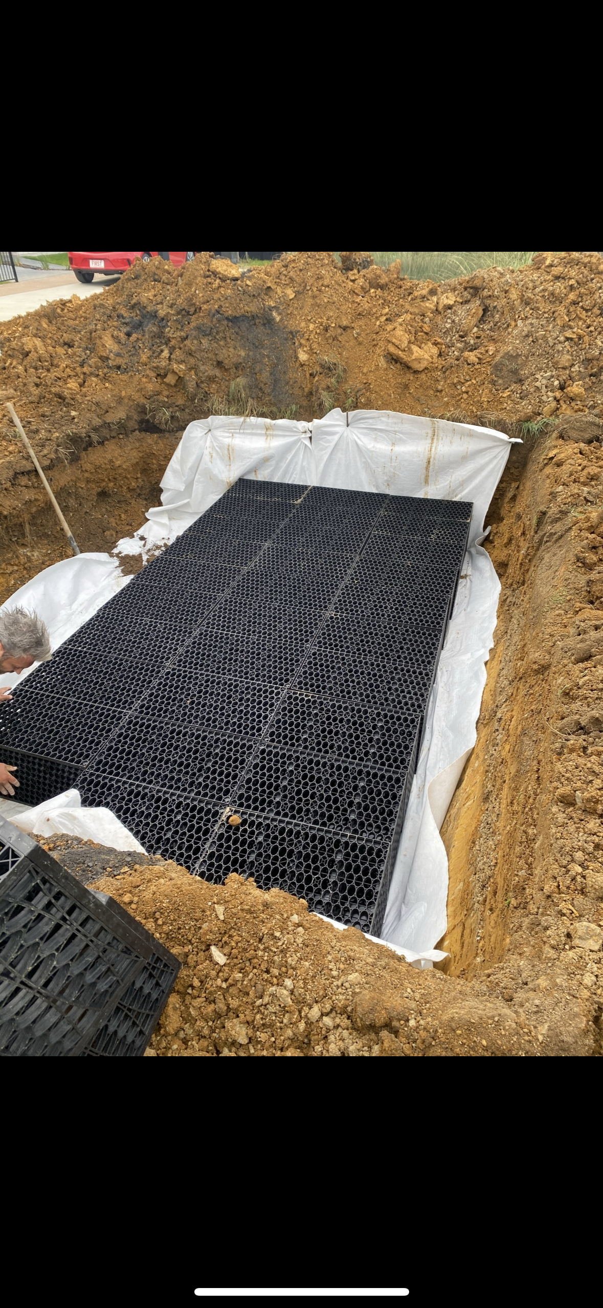 Excavation site with large black plastic grid layer and white lining, surrounded by dirt, with a person working on the left side.
