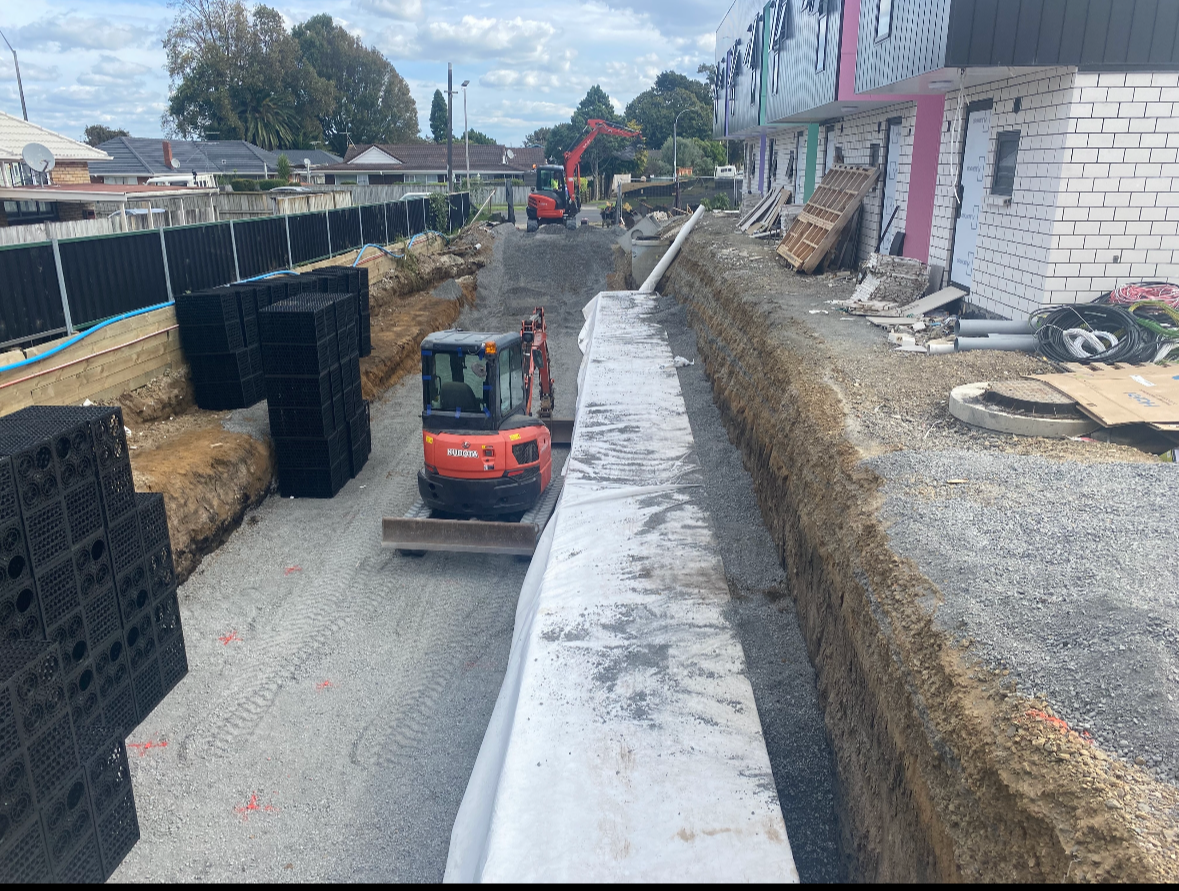 Construction site with excavator working on trench, building foundation, and gravel path, surrounded by construction materials and equipment.