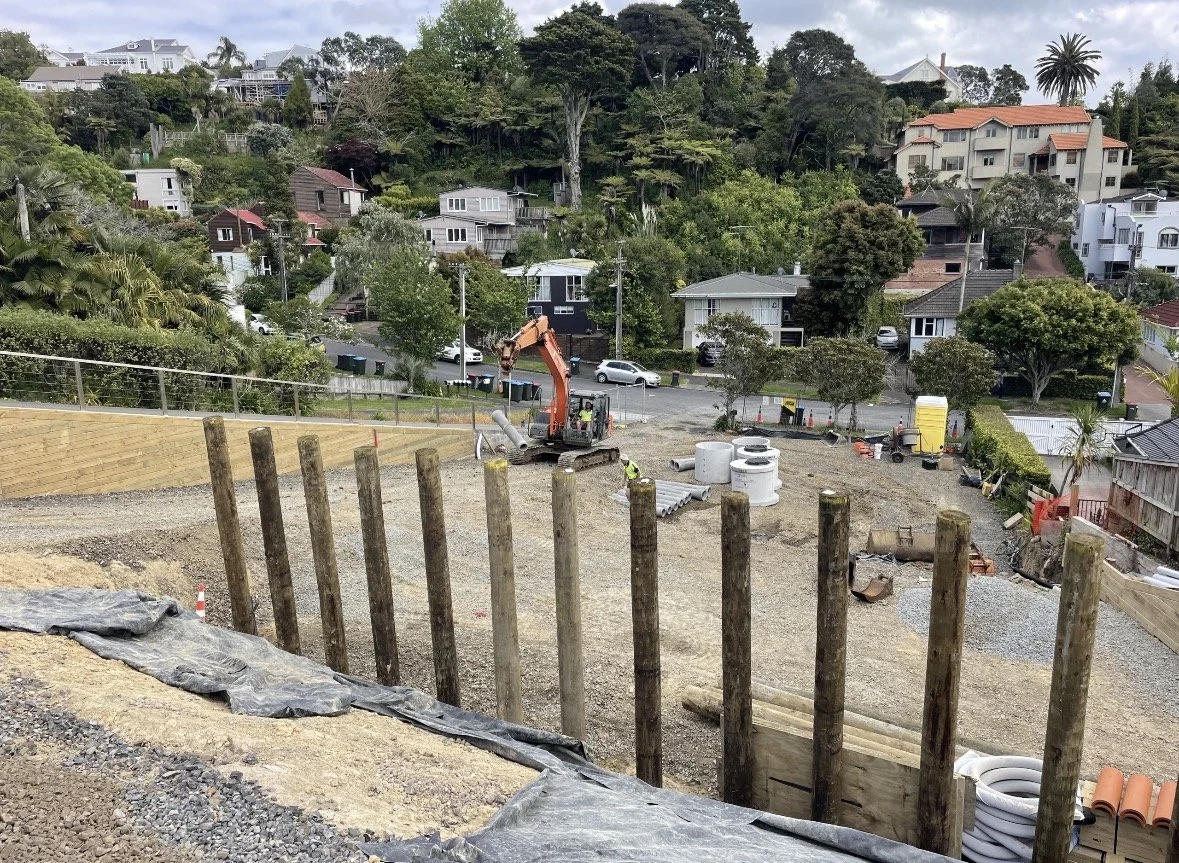Construction site on a hillside with a small excavator and wooden posts, residential houses and trees in the background.