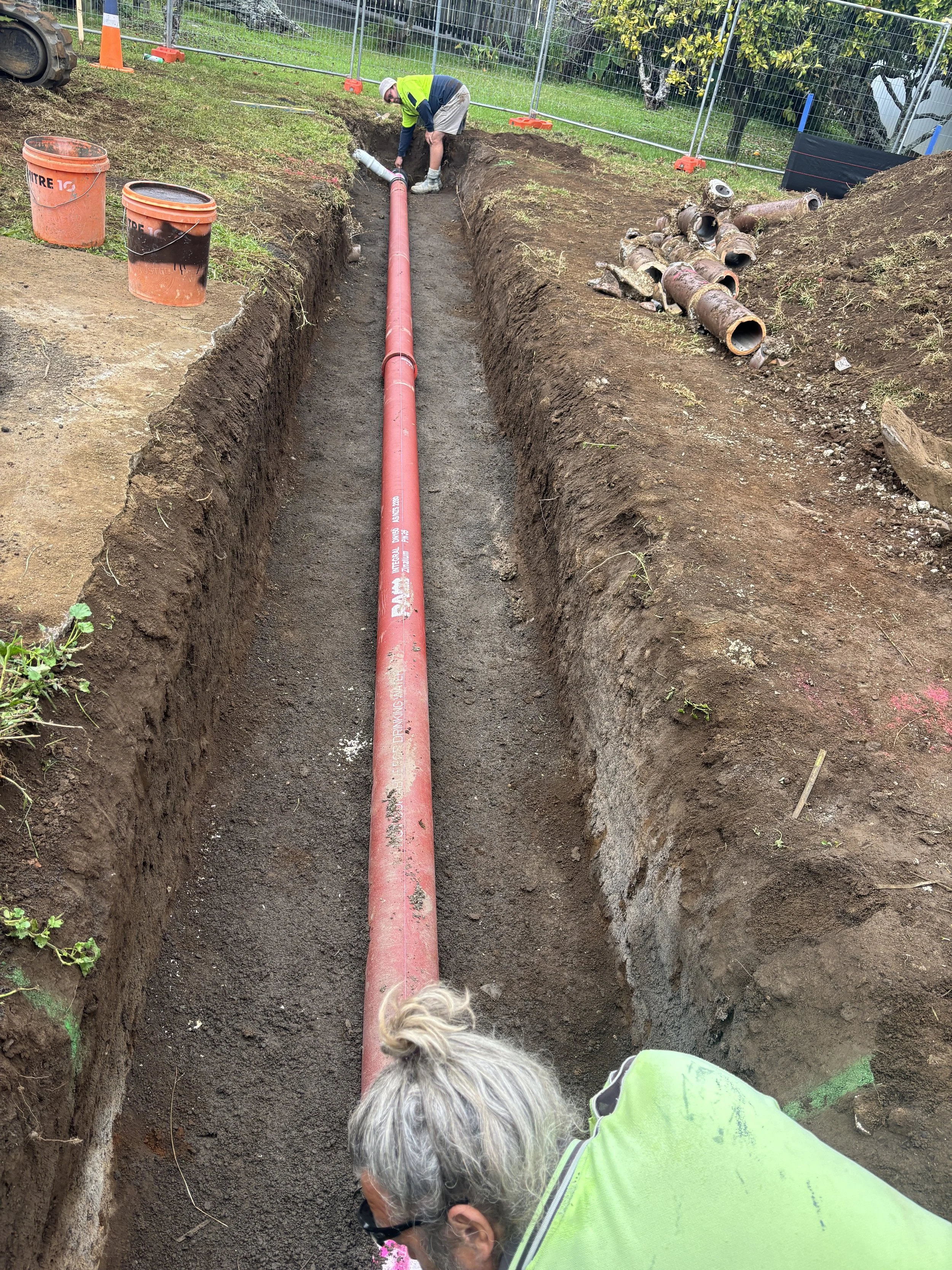 Construction workers installing a red pipe in a trench at a construction site, with tools and orange buckets nearby.
