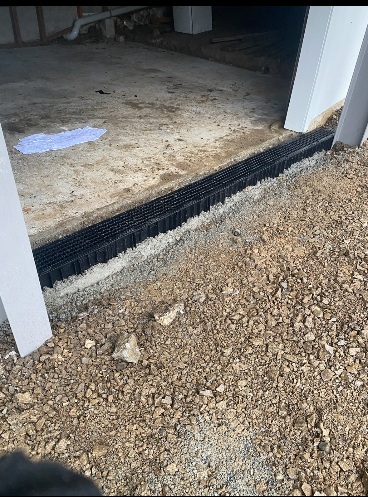 A black plastic drainage grate installed at the entrance of a garage with a gravel driveway outside.