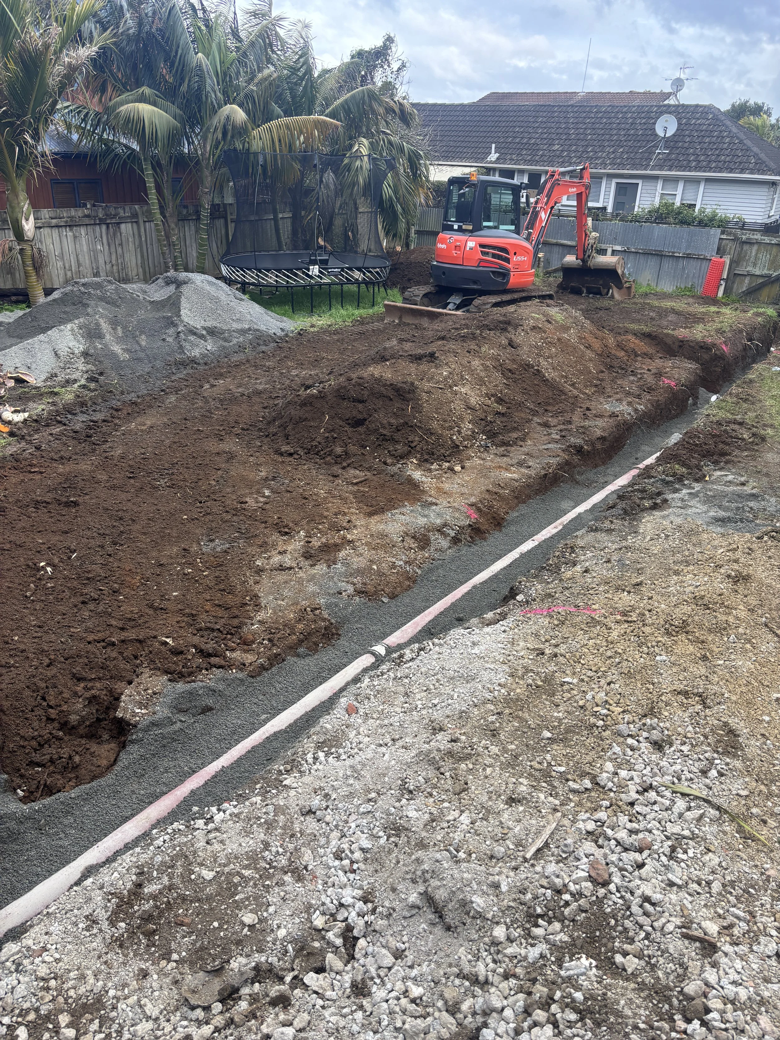 Construction site with a small excavator digging a trench for underground utilities, with gravel and dirt piles, in a backyard with a fenced yard and a trampoline.