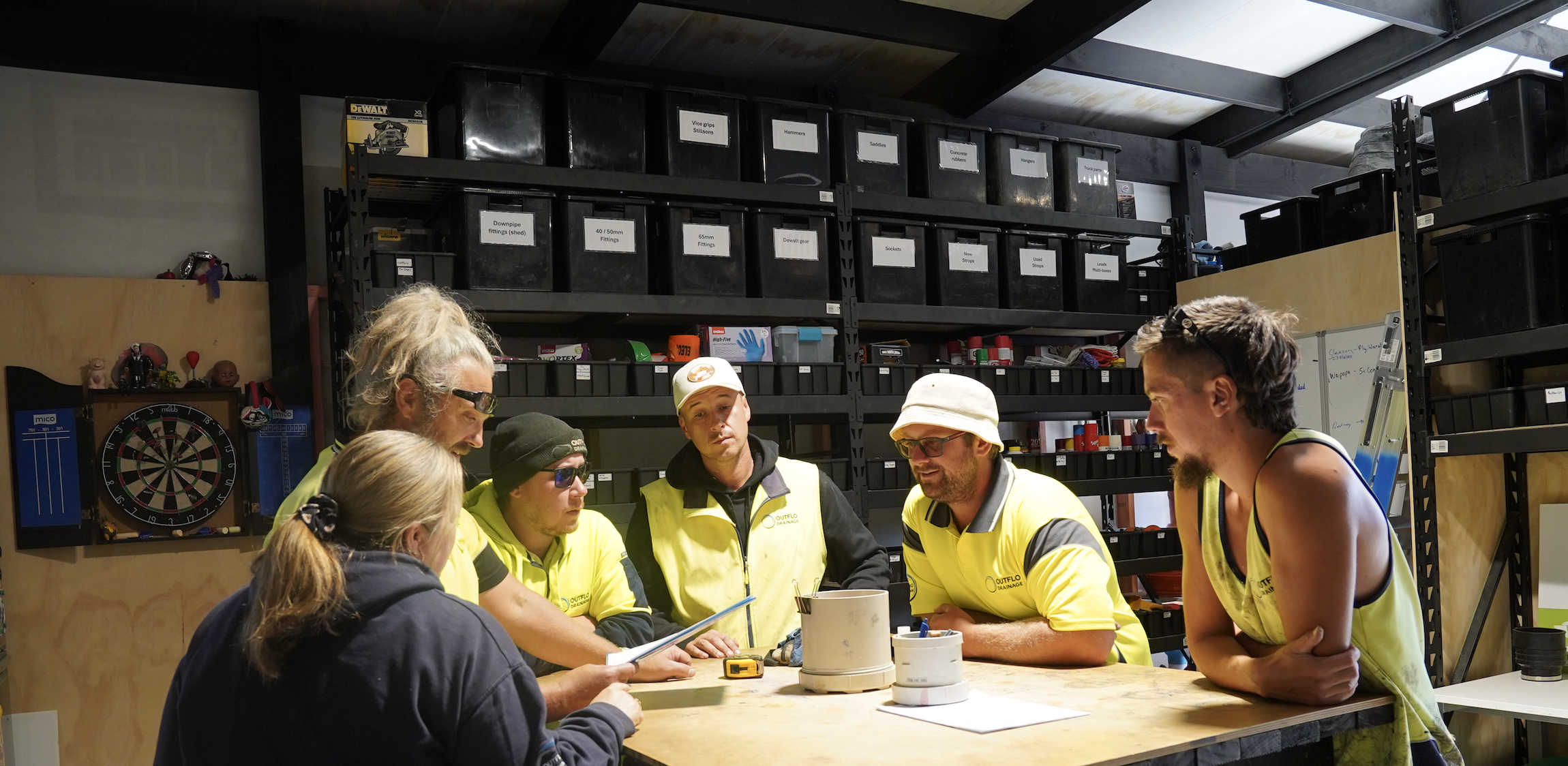 Six people gathered around a work table in a workshop, engaged in a discussion. Behind them are shelves filled with labeled storage bins and tools.