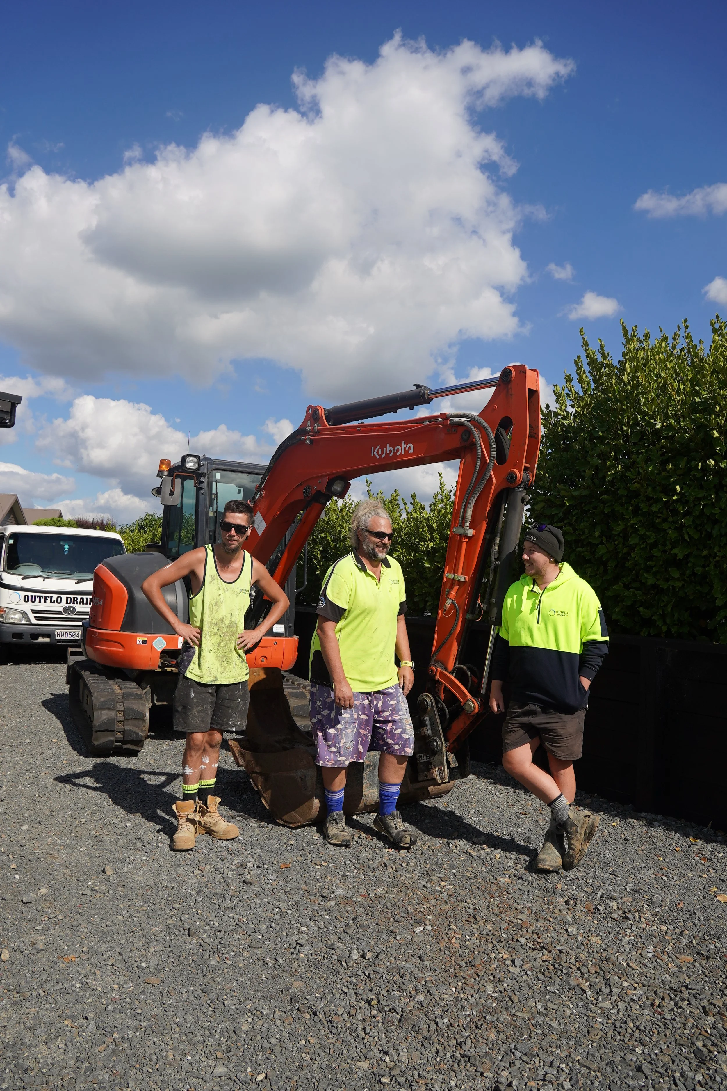 Three construction workers standing in front of a small orange excavator on a gravel surface, with a black fence and green trees behind them, under a partly cloudy sky.