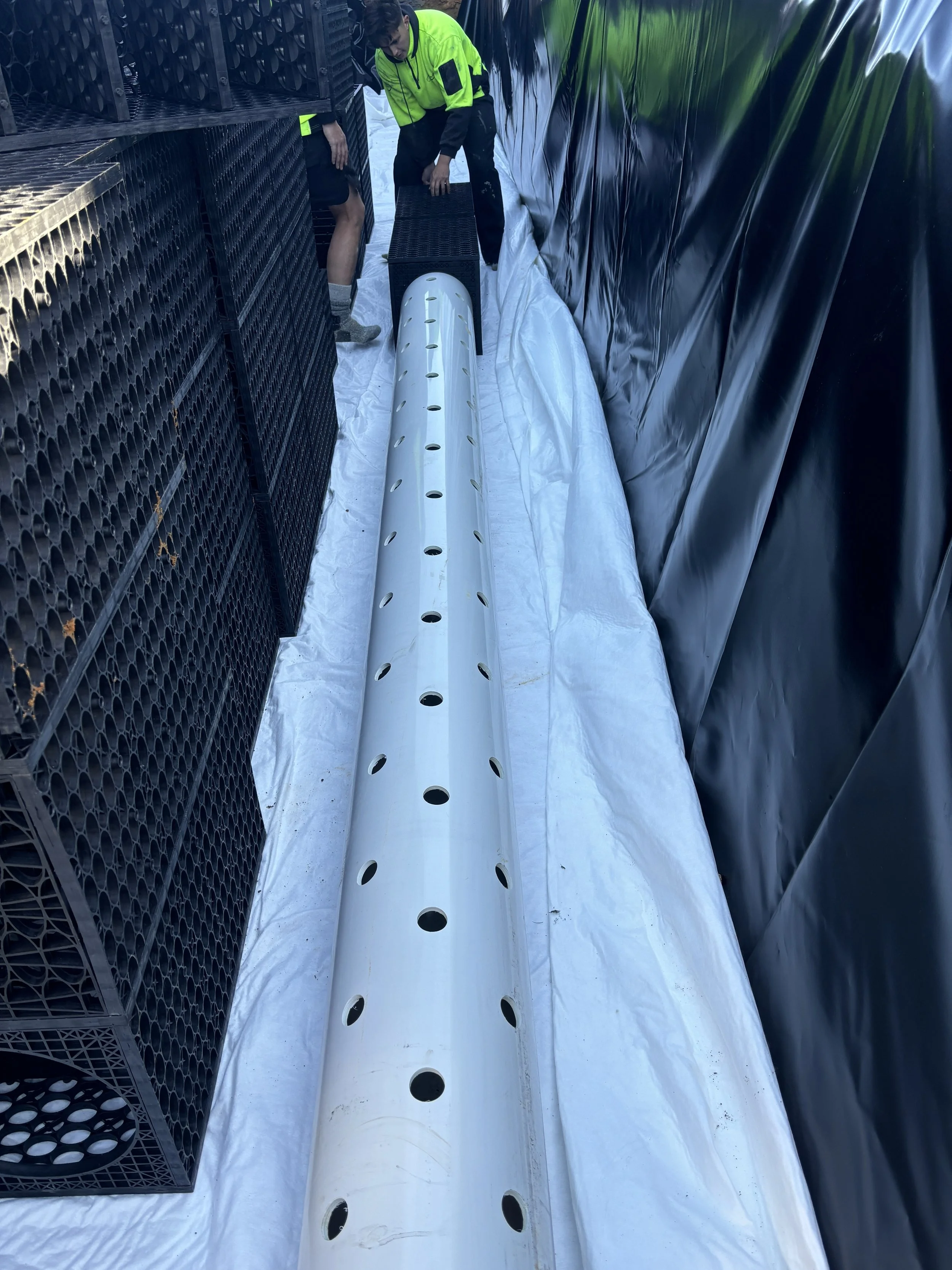 Two workers in safety vests installing or inspecting a large white perforated pipe on a white tarp, surrounded by black plastic sheeting and black crates.
