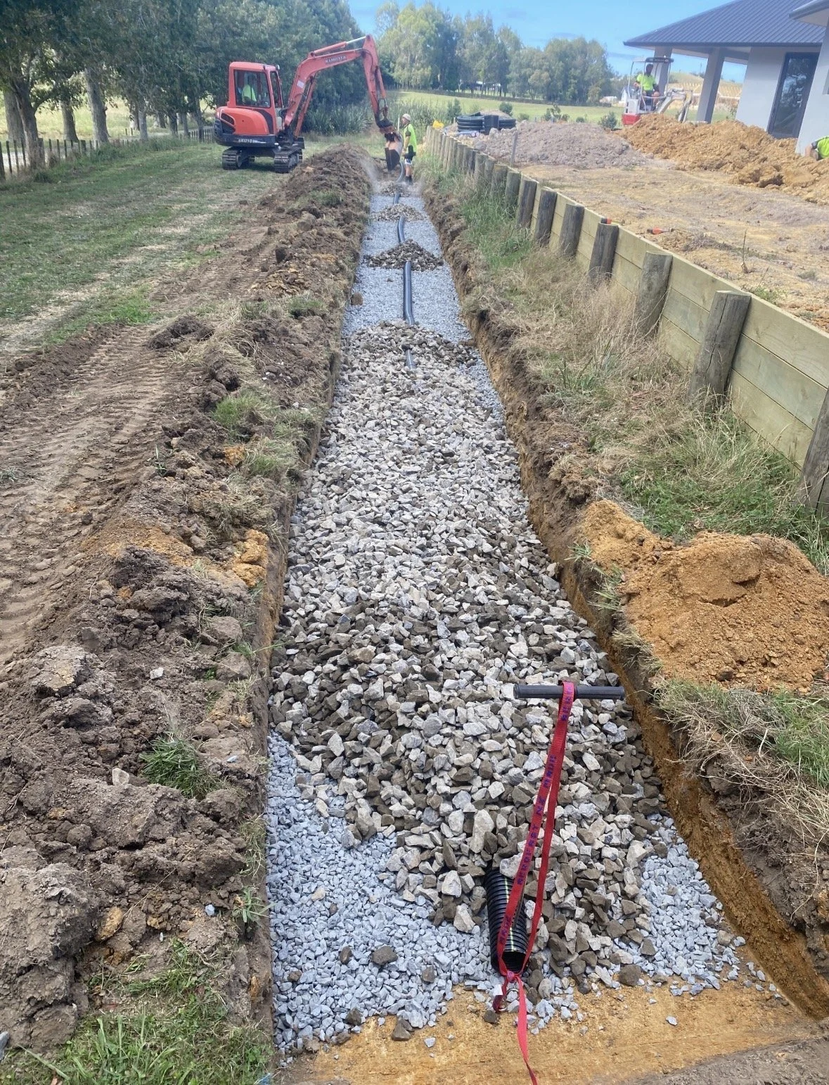 Construction workers building a gravel drainage trench with a mini excavator, surrounding a new house in a rural area.