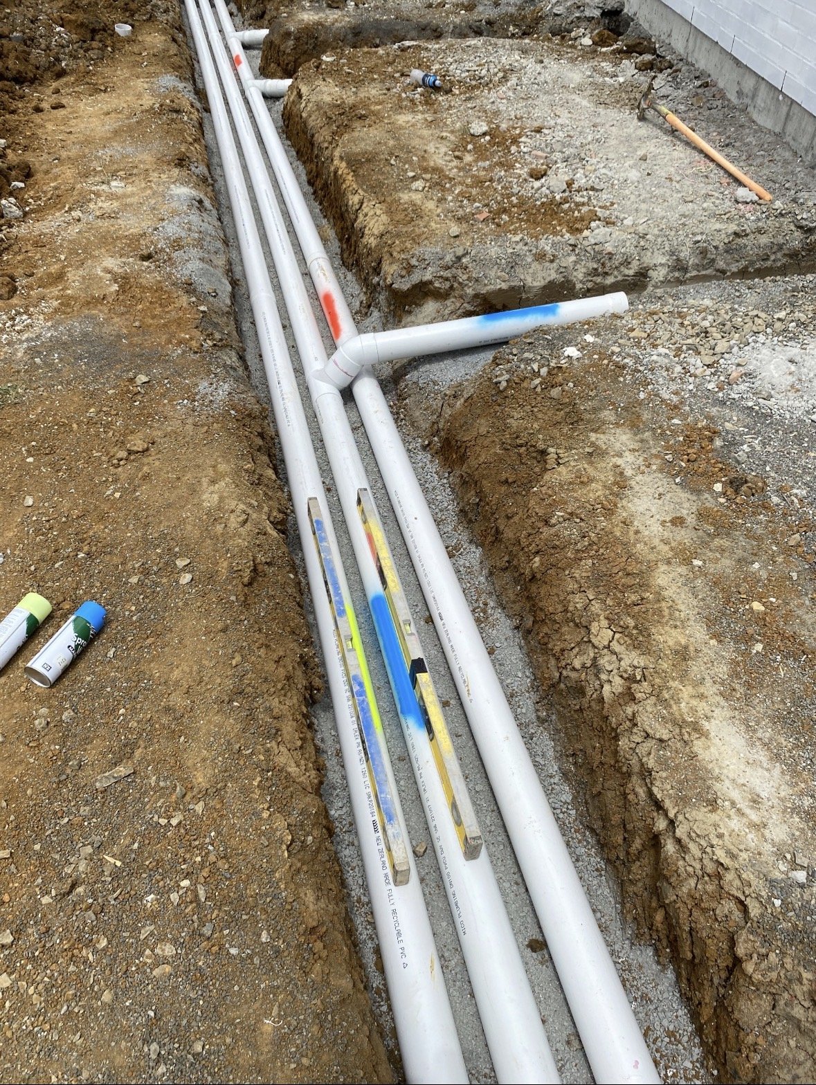 Construction site with exposed white PVC pipes in a trench, some with colored markings, near a building with a white exterior wall.