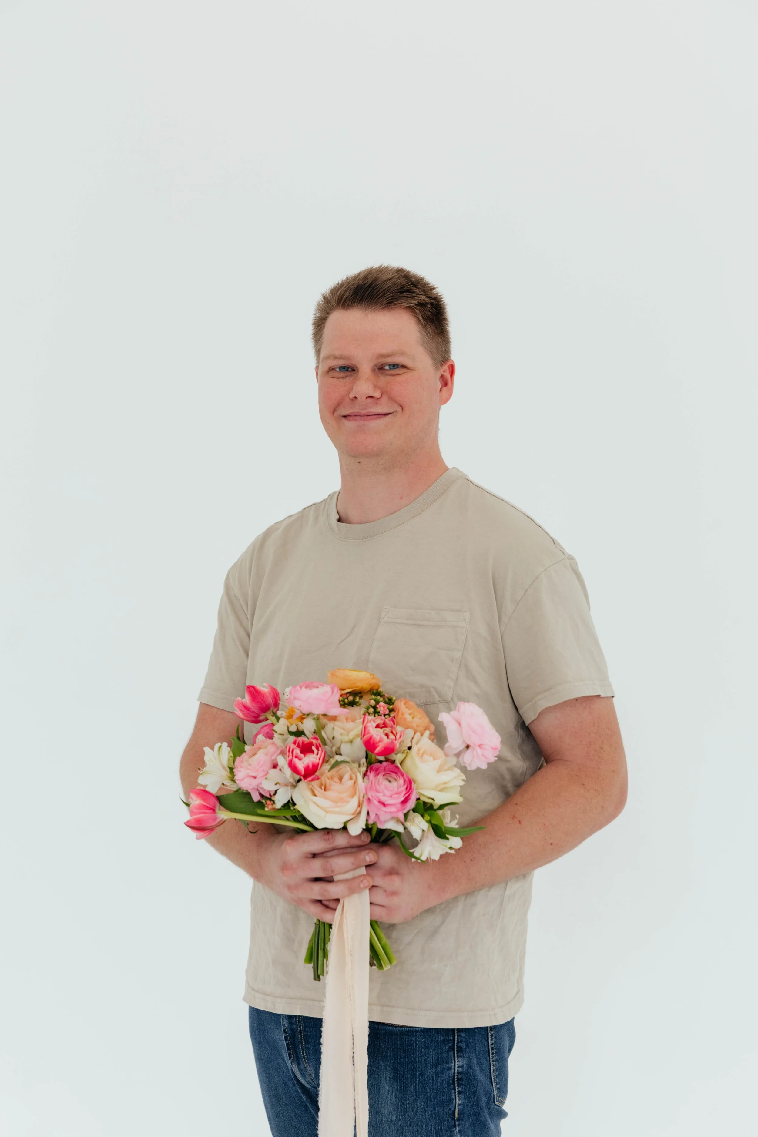 A man with short brown hair and a light beige t-shirt holding a large bouquet of pink, white, and peach flowers.