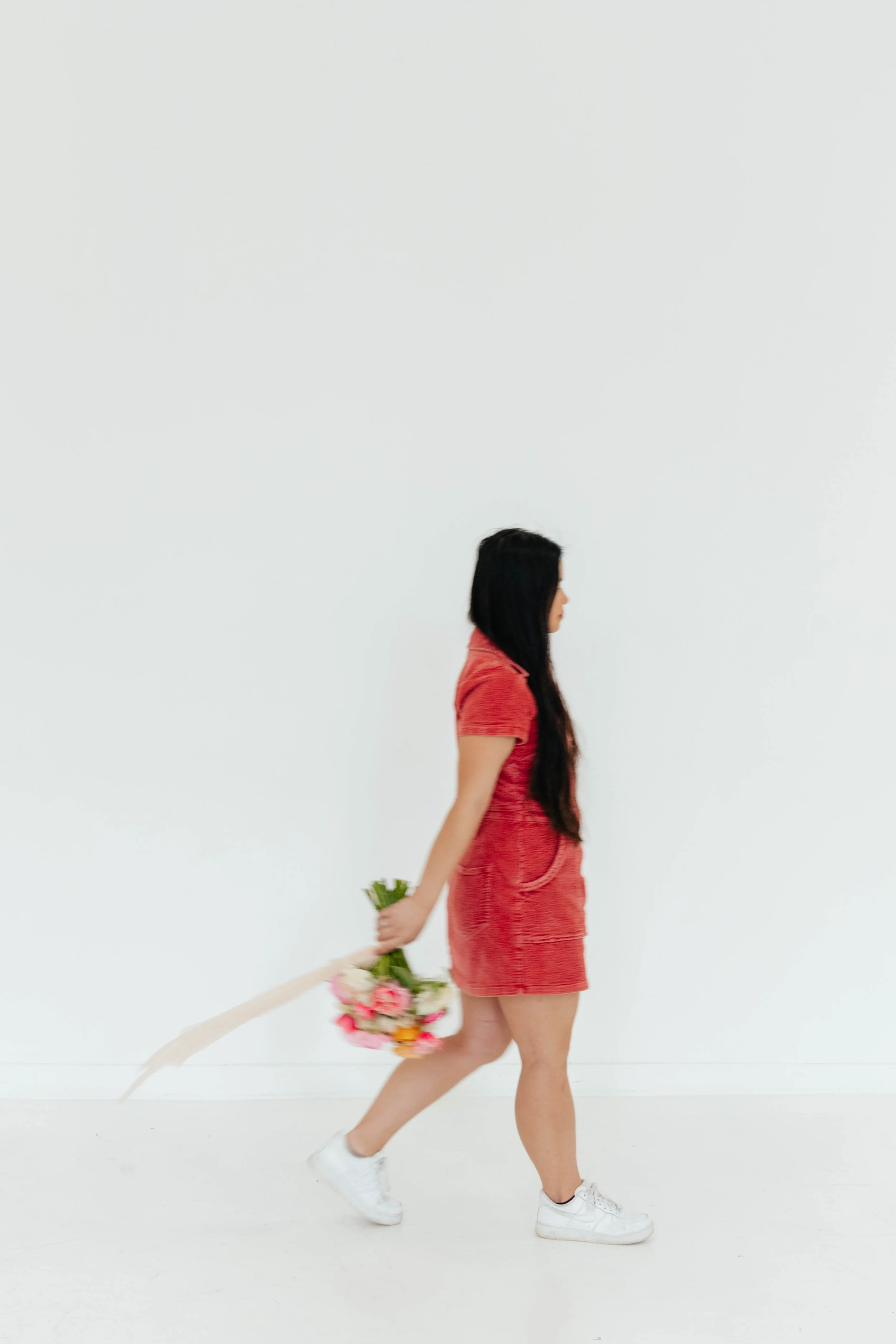 A woman in a red dress walking, holding a bouquet of flowers behind her back.