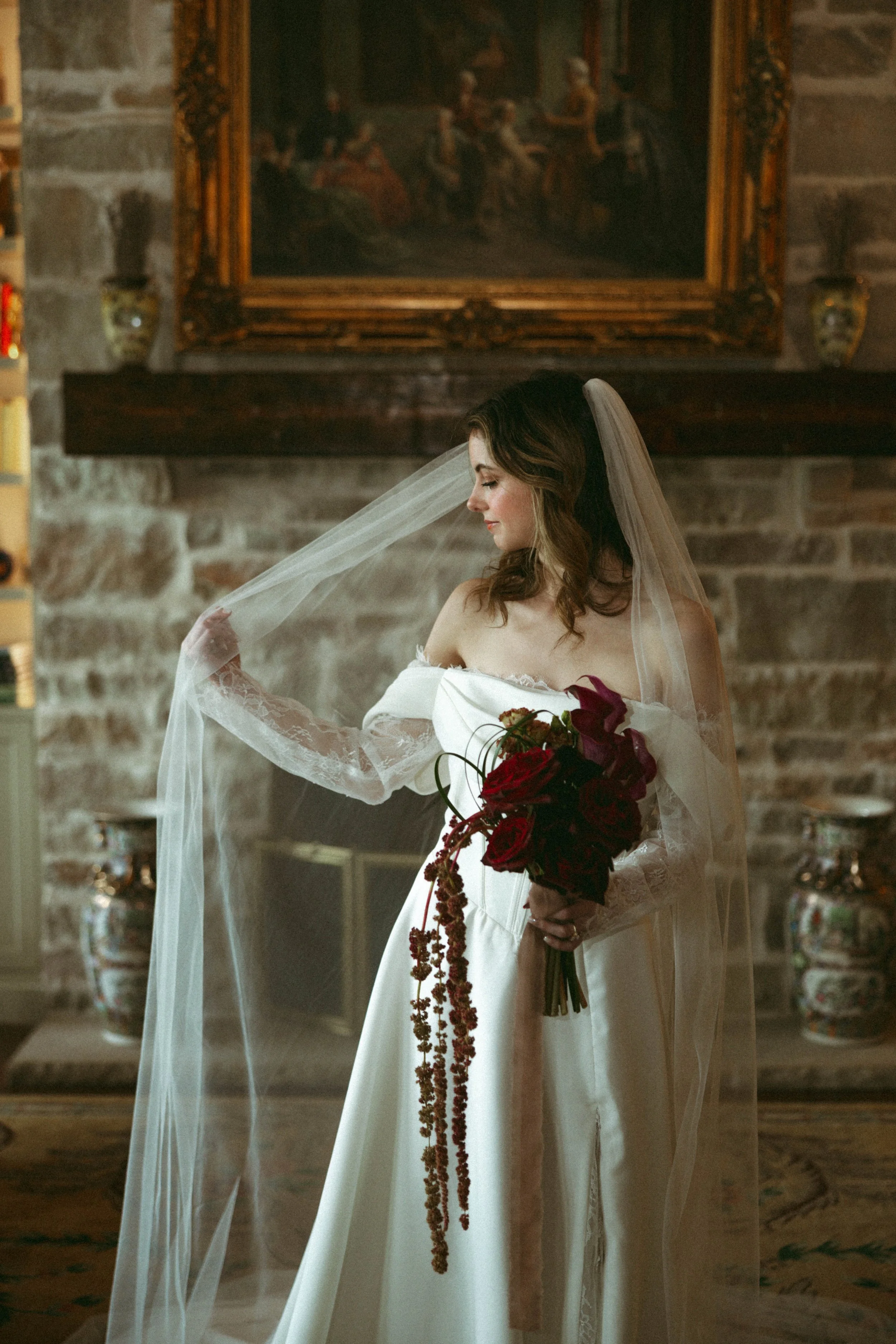 A bride with light skin and brown hair holding a bouquet of red flowers and wearing a long veil.
