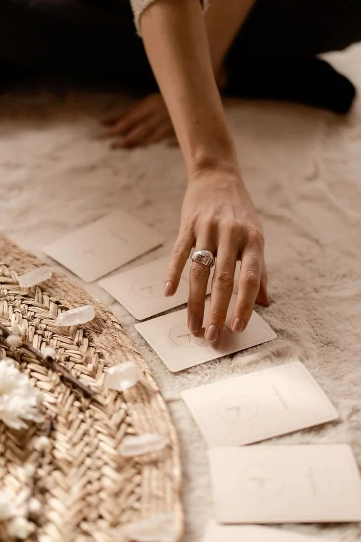 A person's hand with rings placed on tarot cards on a soft surface, with some cards face down and a woven basket in the foreground.