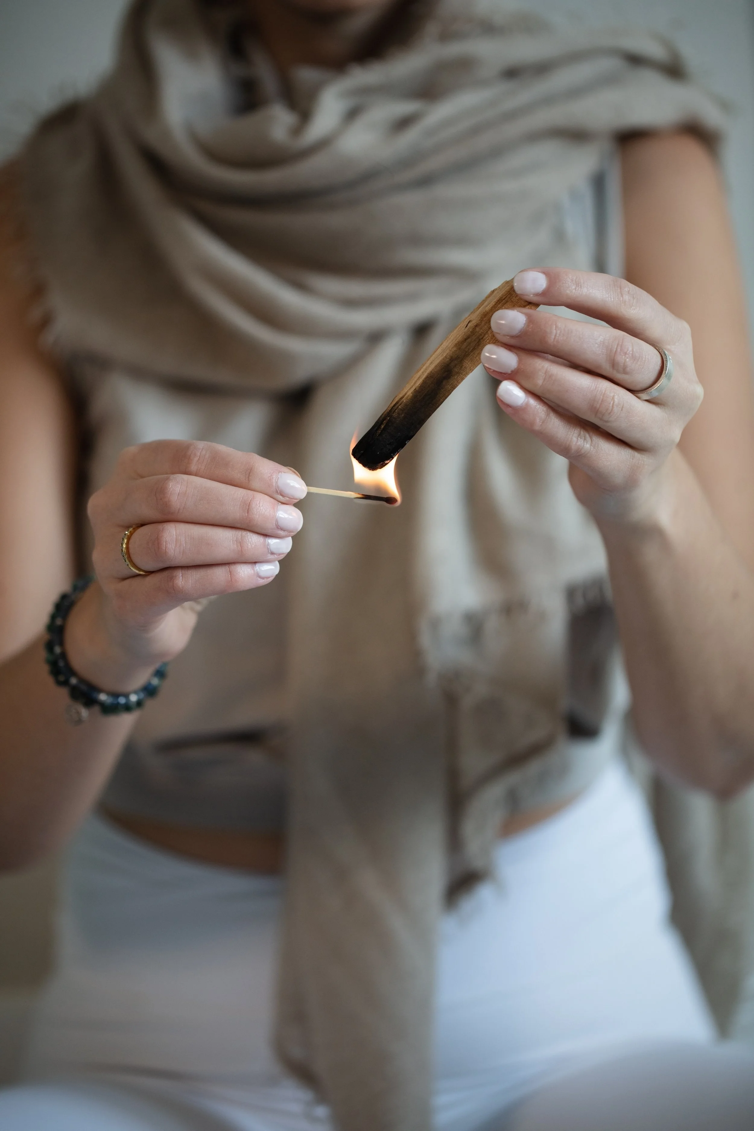 A person holding a lit wooden matchstick with flames, wearing a beige scarf, ring, and bracelet, with a blurred background.
