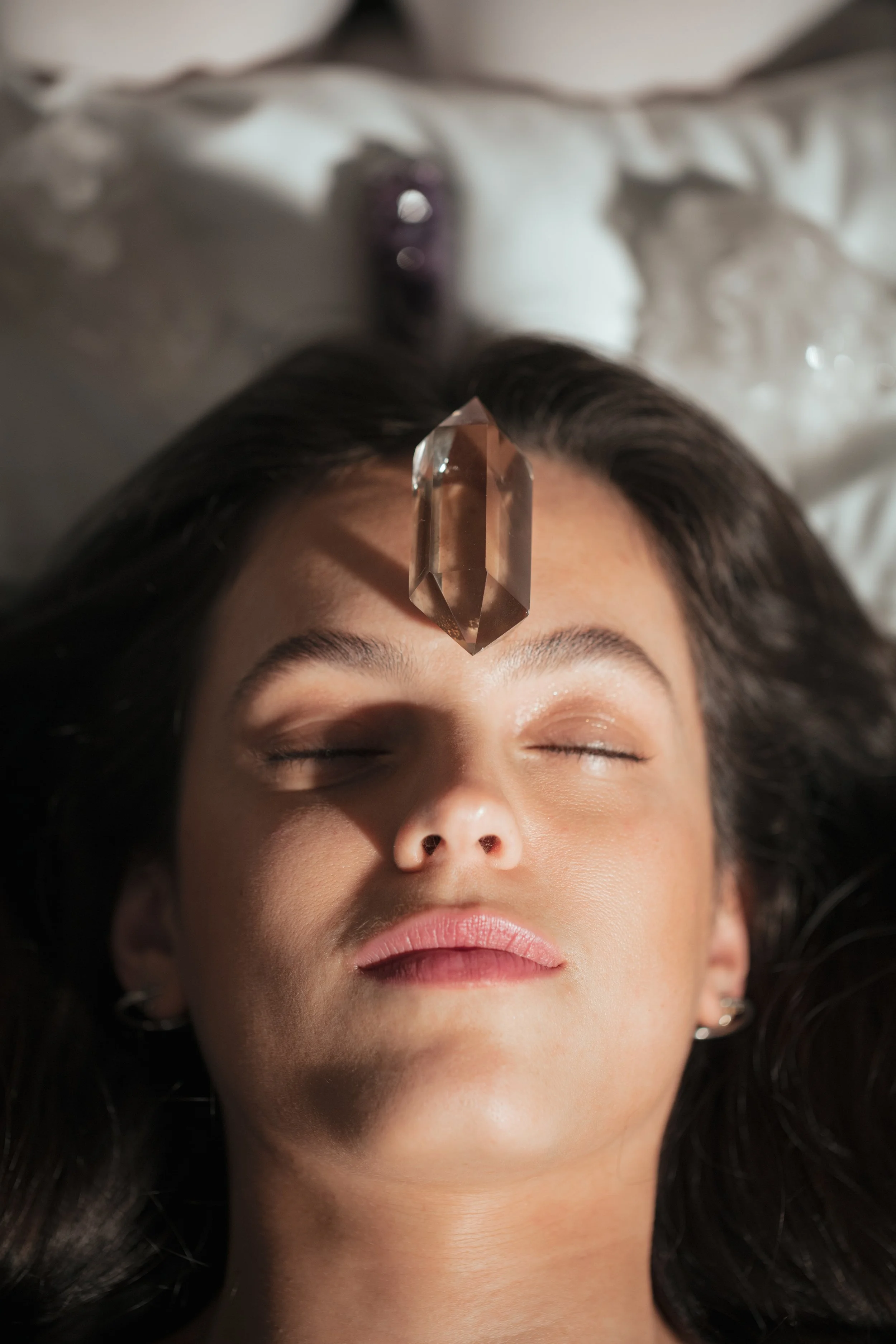 A woman lying on her back with her eyes closed, a large crystal on her forehead, and dark hair spread around her head.