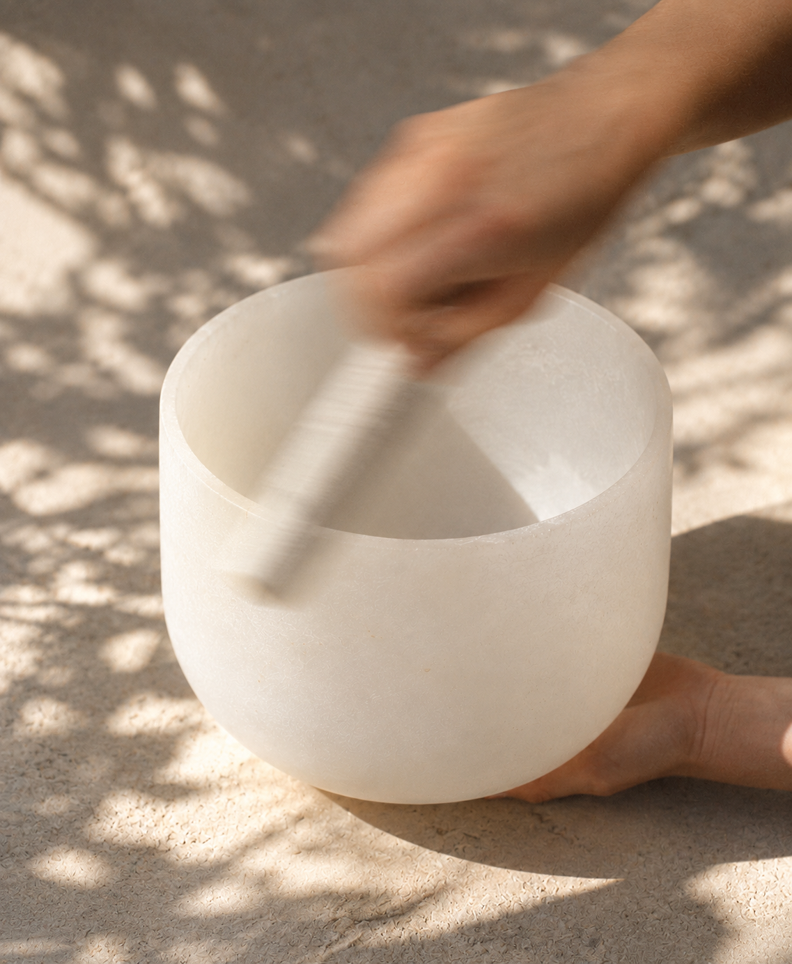Hand playing a white crystal singing bowl in dappled sunlight