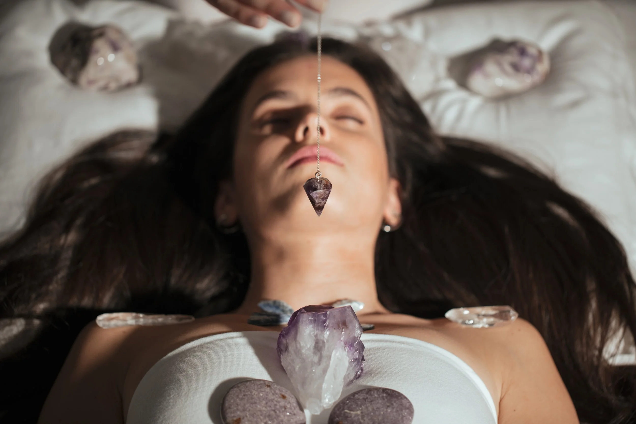 A woman lying down with her eyes closed, surrounded by various large crystals and stones, with one crystal hanging from a chain above her face.