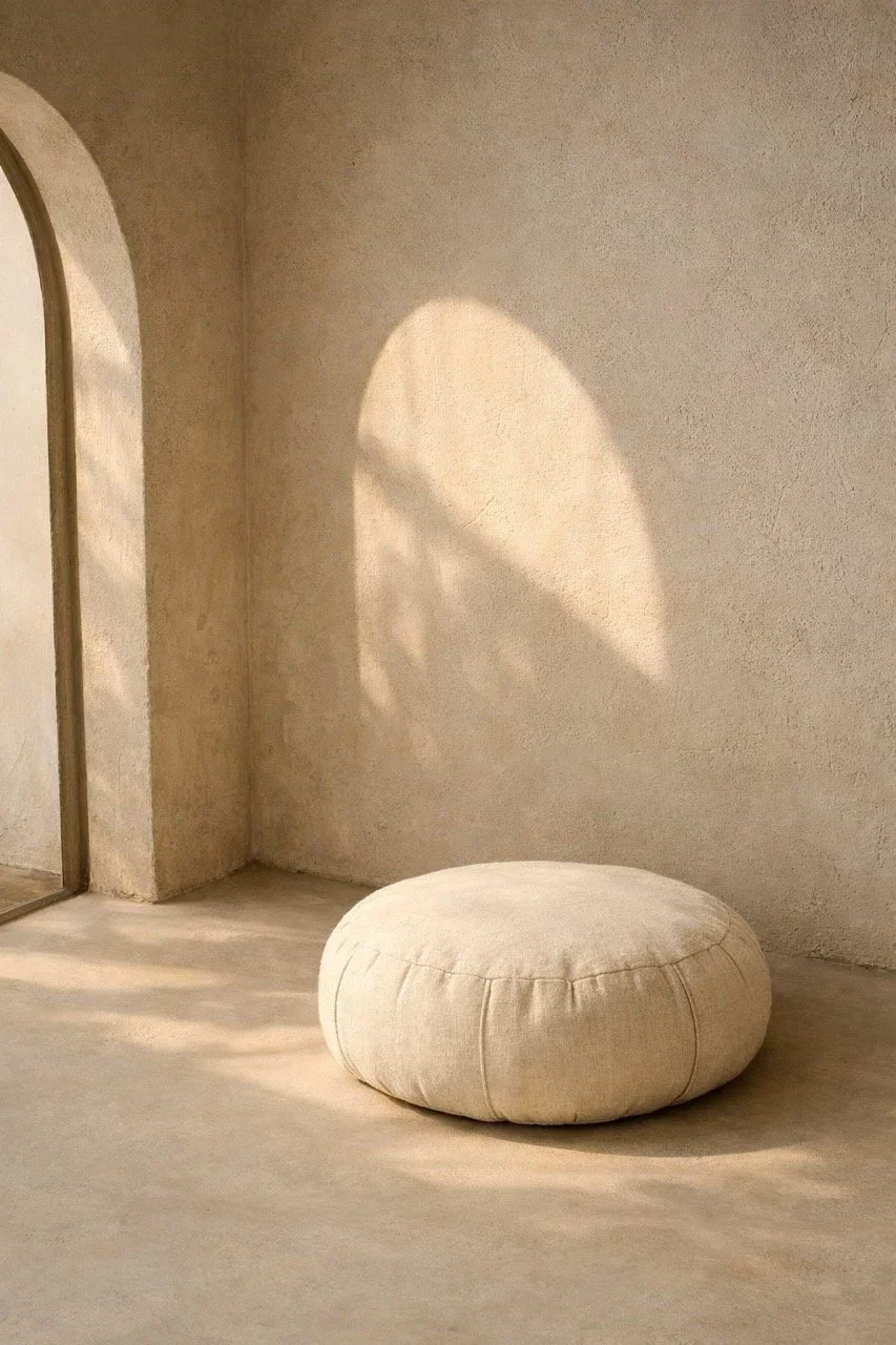 A round, beige, fabric ottoman on a light-colored floor, with a soft shadow of a circular window or light fixture cast on the wall behind it.