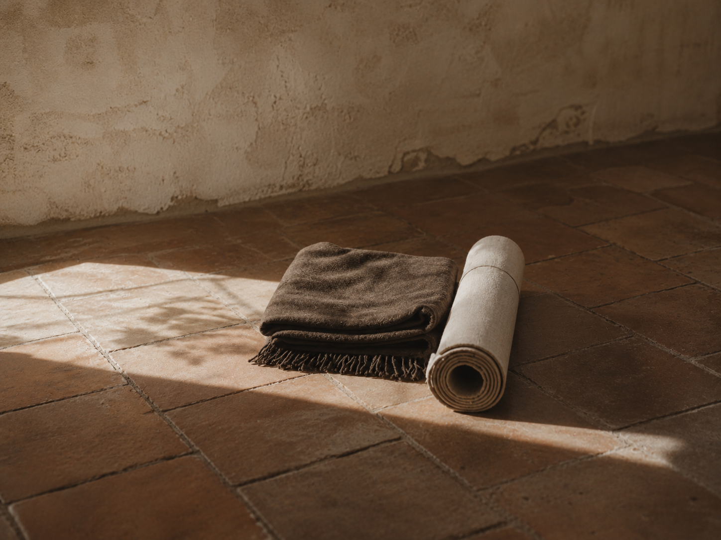 A rolled-up beige rug and a folded dark brown blanket are placed on a terracotta tiled floor near a textured wall, with sunlight casting shadows across the scene.