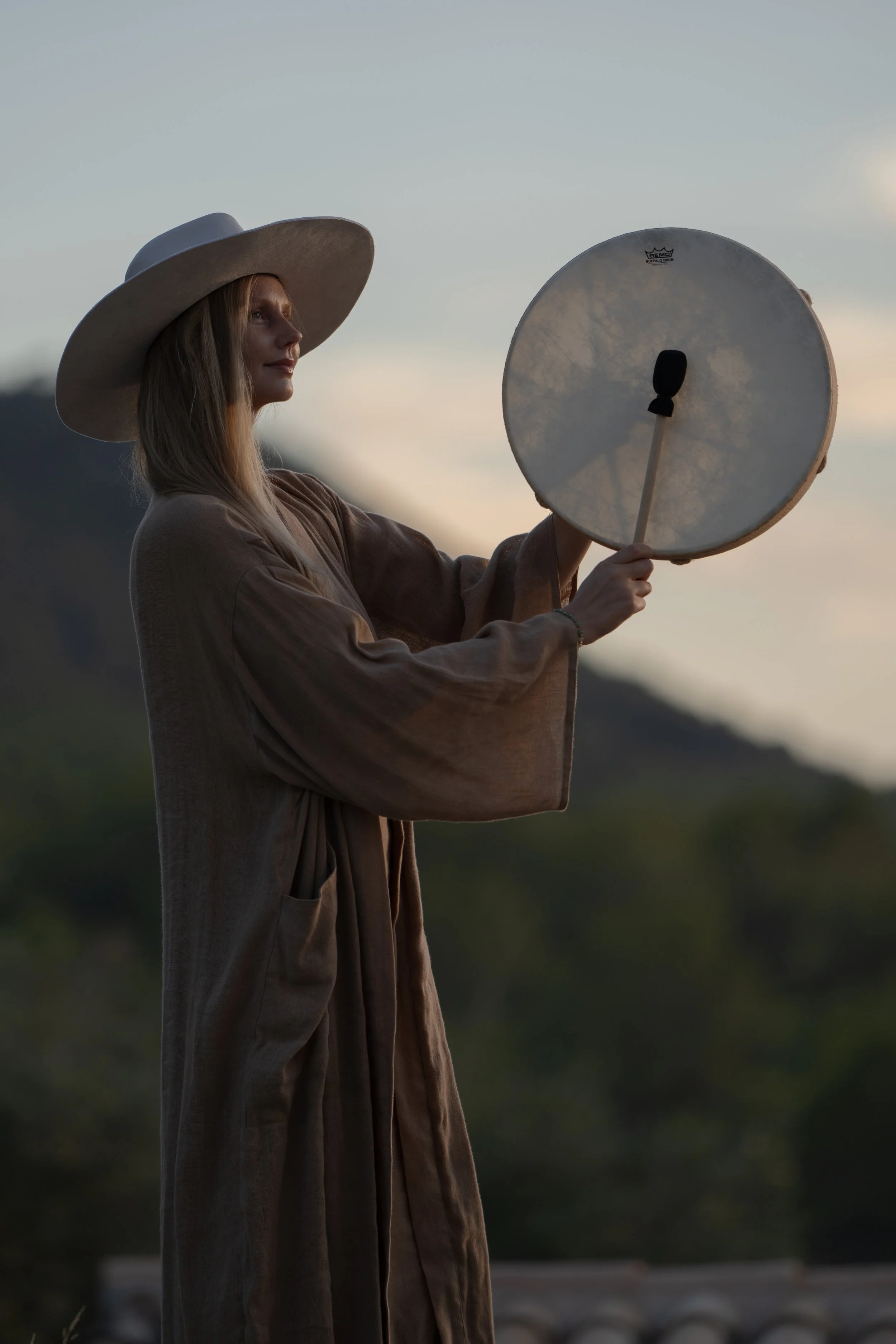 A woman wearing a large sunhat and a brown dress holding a round drum outdoors during sunset.