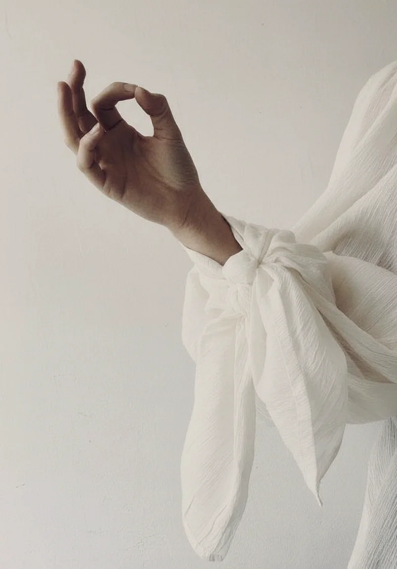 A person making an OK sign with their hand, wearing a white long-sleeve shirt with a tied cuff, against a plain light-colored wall.