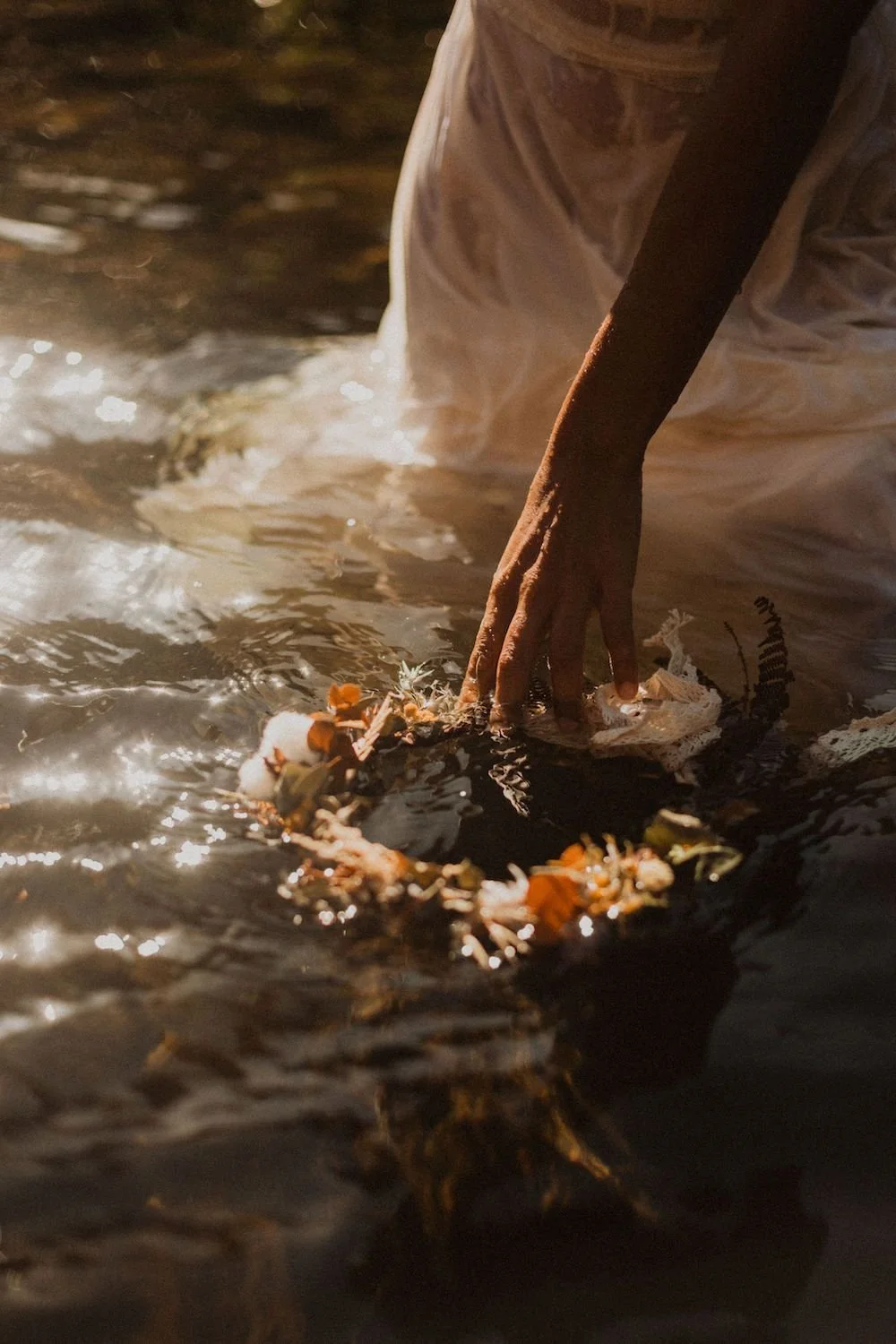 A person wearing a white garment is kneeling by a body of water at night, placing a flower wreath into the water. The person's hand is visible, and the scene is softly illuminated, with petal petals and leaves floating on the water's surface.