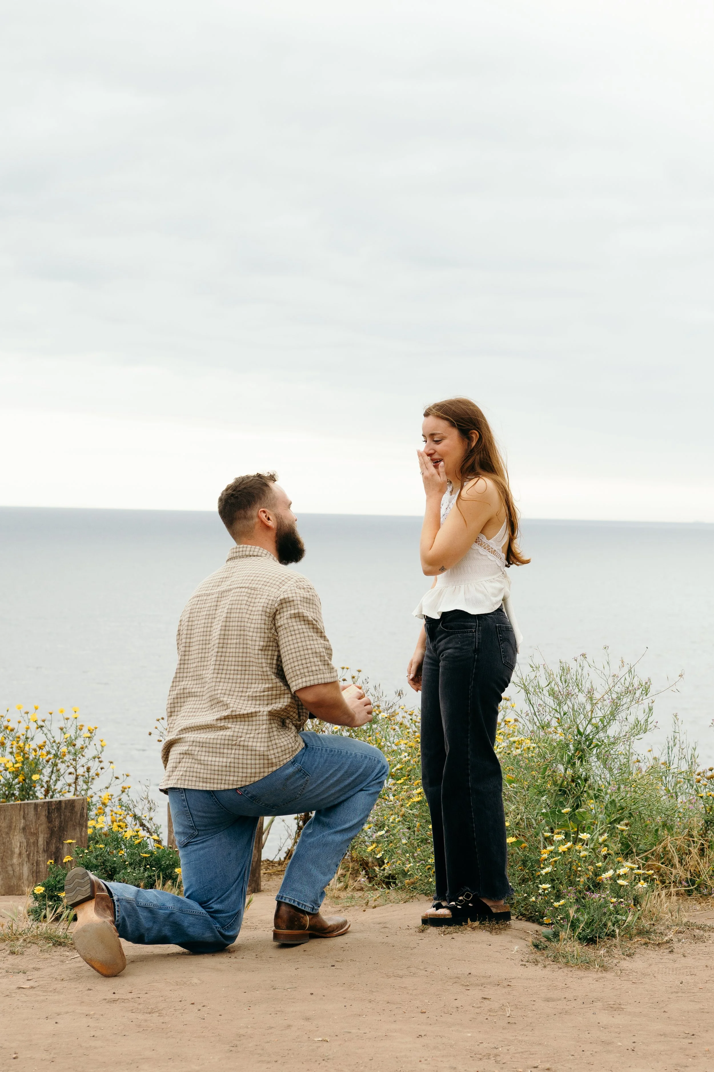A man on one knee proposing marriage to a woman standing outdoors near the ocean. The woman is smiling and covering her mouth with her hand, and there are flowers and greenery around them.