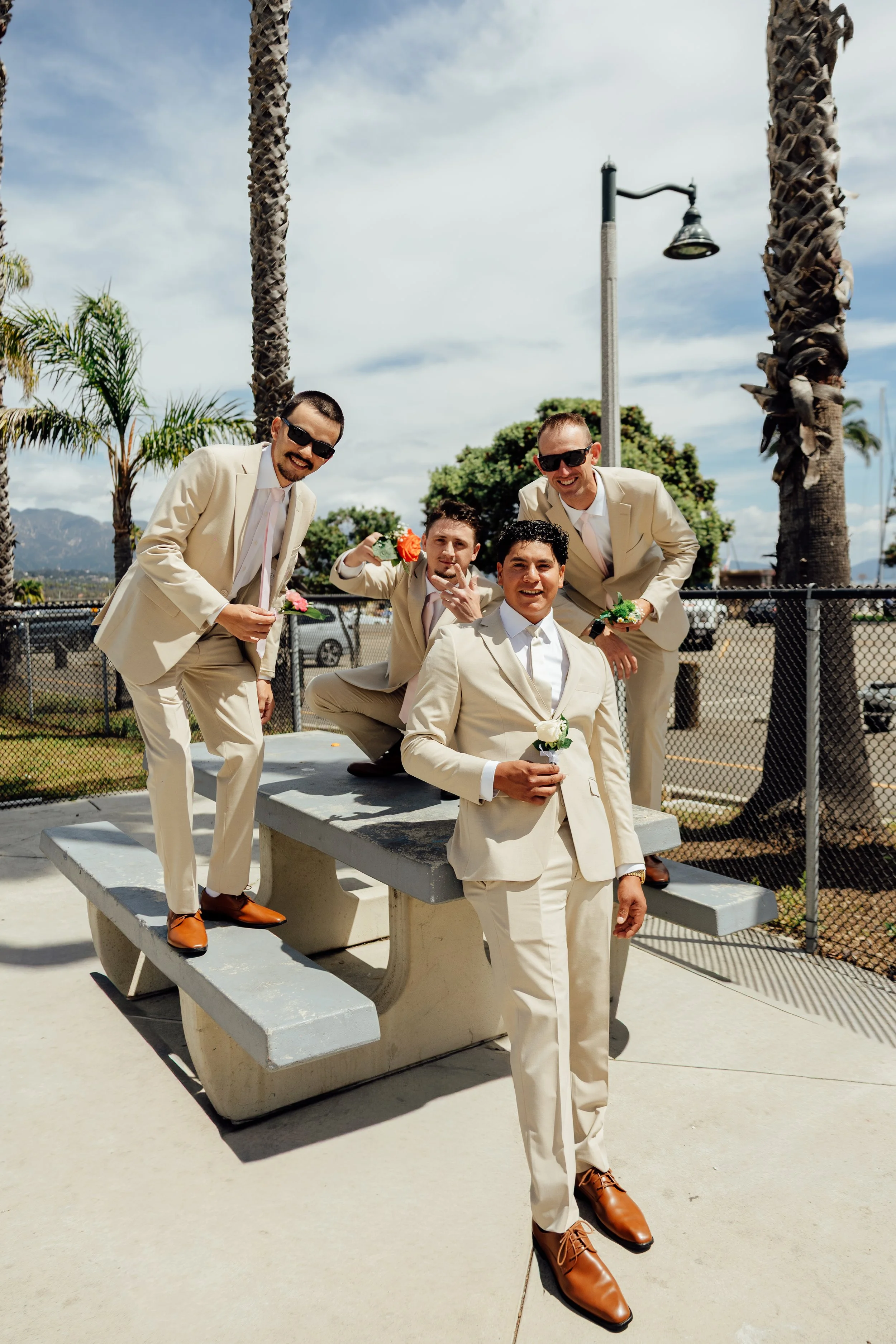 Four men in beige suits and dark sunglasses posing outdoors with palm trees, a fence, and mountains in the background on a partly cloudy day.