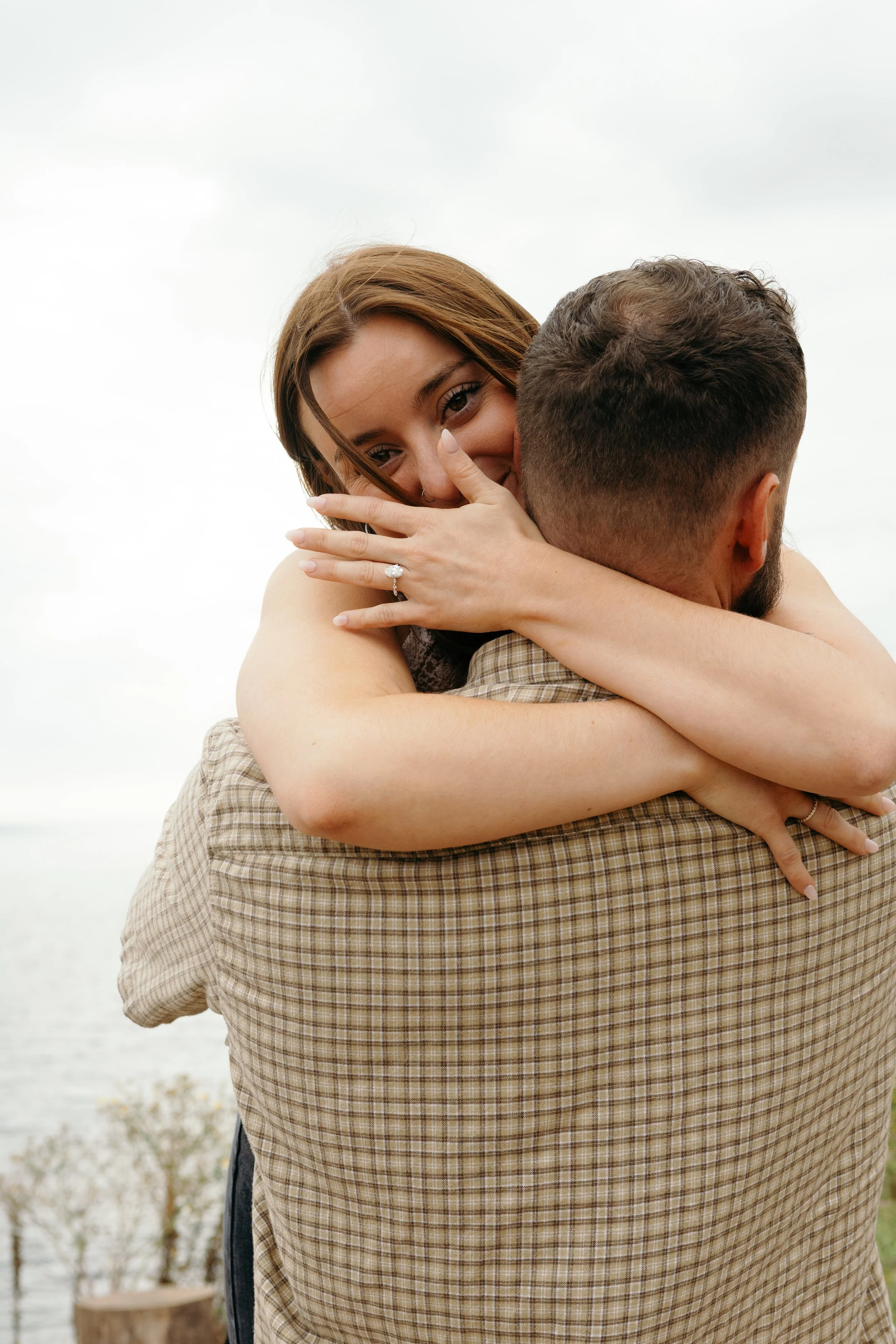 A woman hugging a man outdoors, with her arms around his shoulders, showing an engagement ring on her finger, and smiling at the camera.