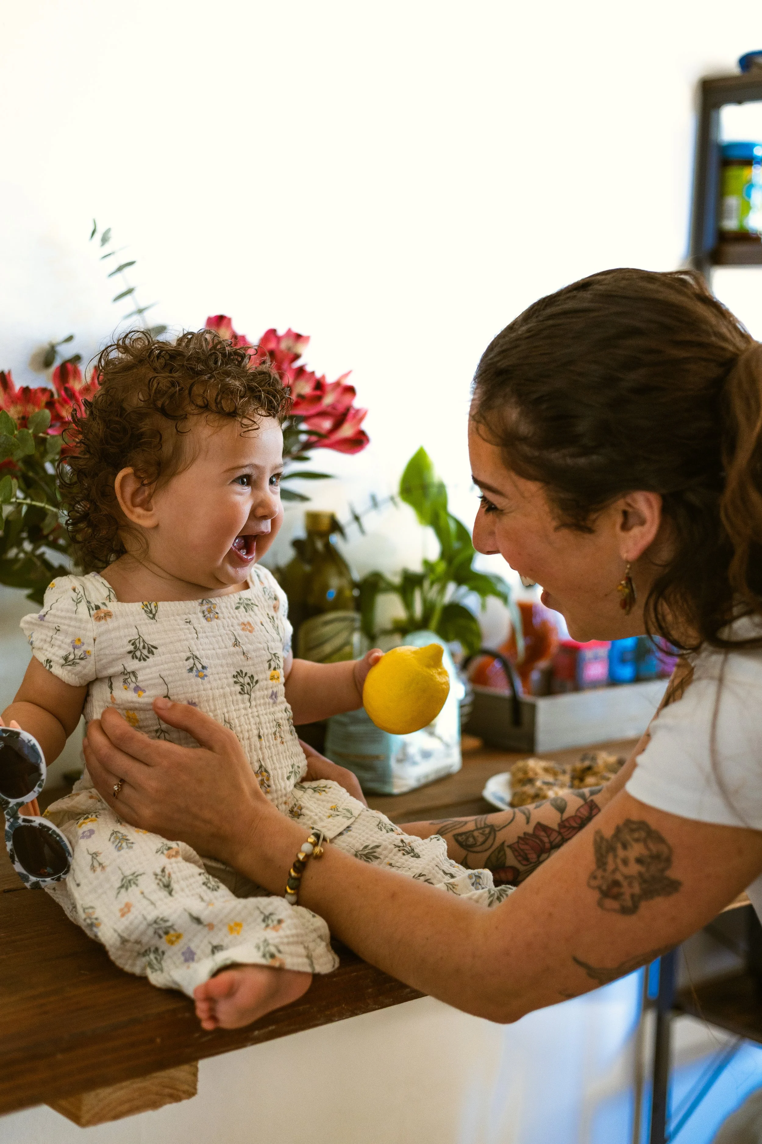 A woman and a young girl are smiling and laughing at each other in a kitchen. The girl is holding a large yellow lemon, and there are flowers and kitchen items in the background. An in home photoshoot in Santa Barbara, CA.
